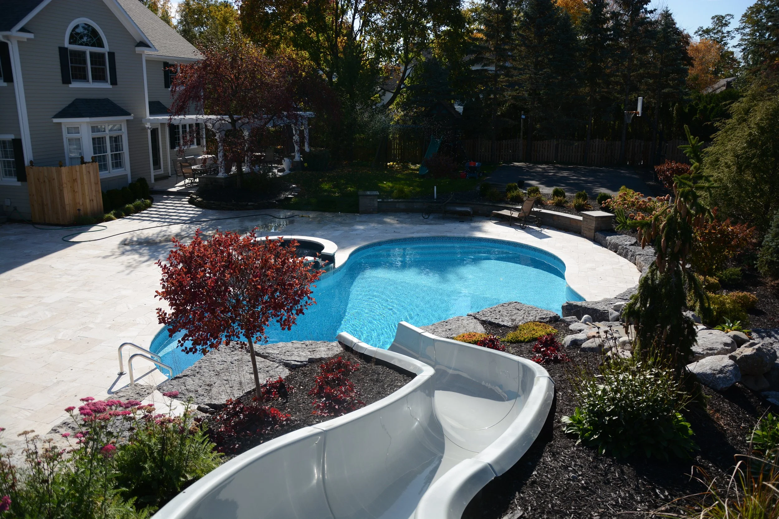 Backyard with a swimming pool, hot tub, and water slide surrounded by plants, trees, and outdoor furniture.