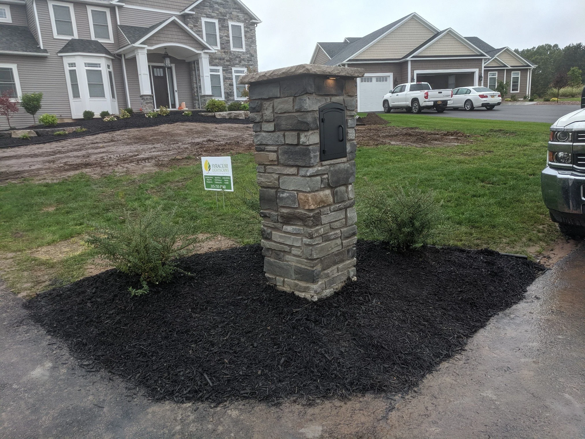 Stone mailbox with a black mailbox door, surrounded by a freshly mulched flower bed with small bushes, in front of a large house with gray siding and a smaller house with a garage in the background, on a rainy day.