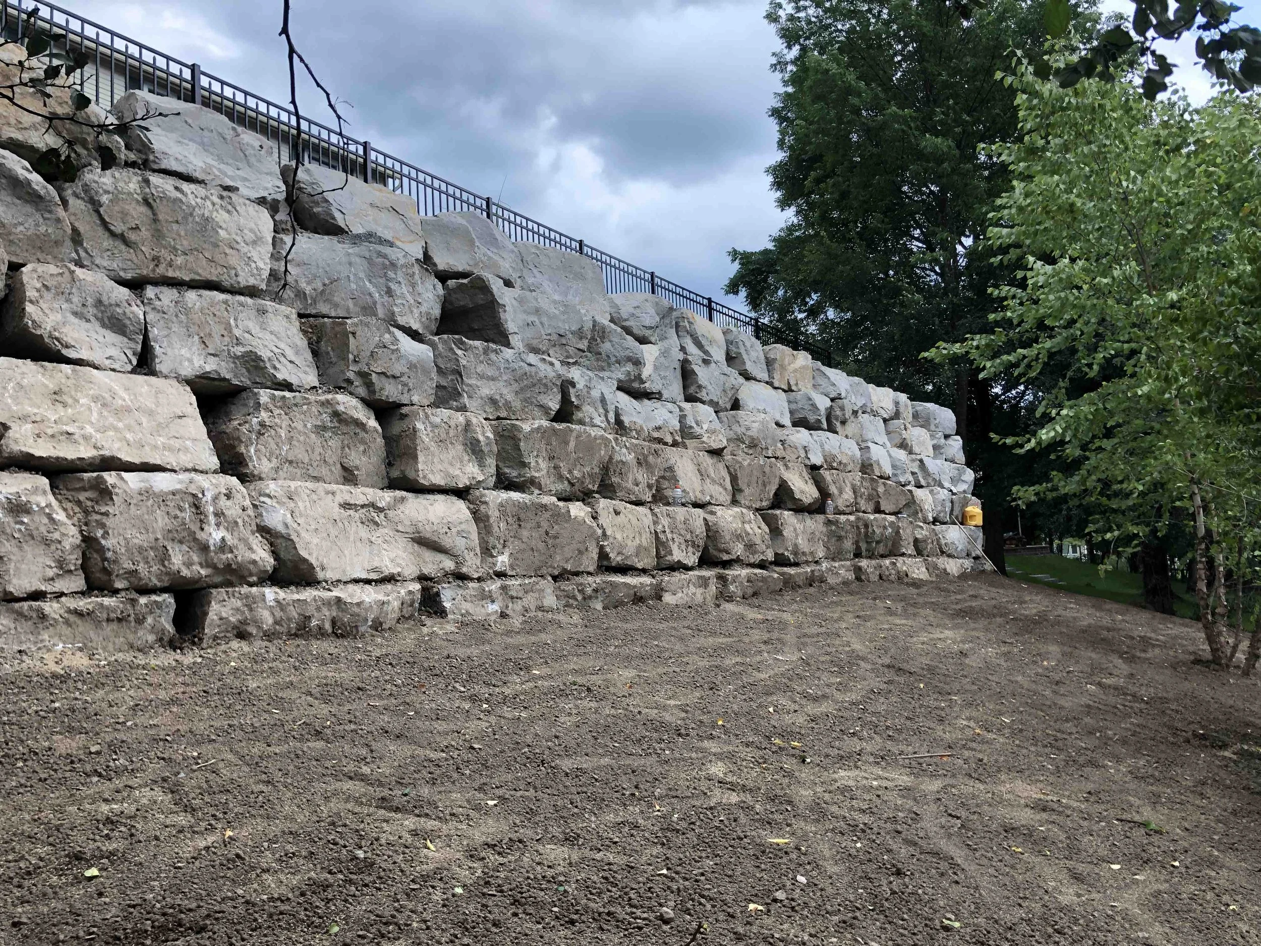 A large stone retaining wall with a metal fence on top, adjacent to a dirt ground and green trees in the background under a cloudy sky.