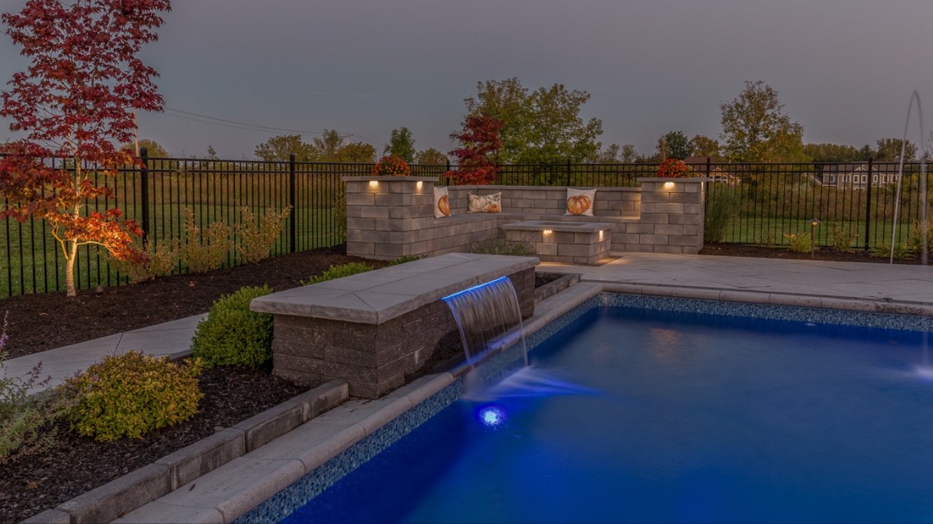 Backyard with a swimming pool, water feature with a waterfall, outdoor seating area with pillows, landscaped garden with trees, flower pots, and landscape lighting, enclosed by a black metal fence.