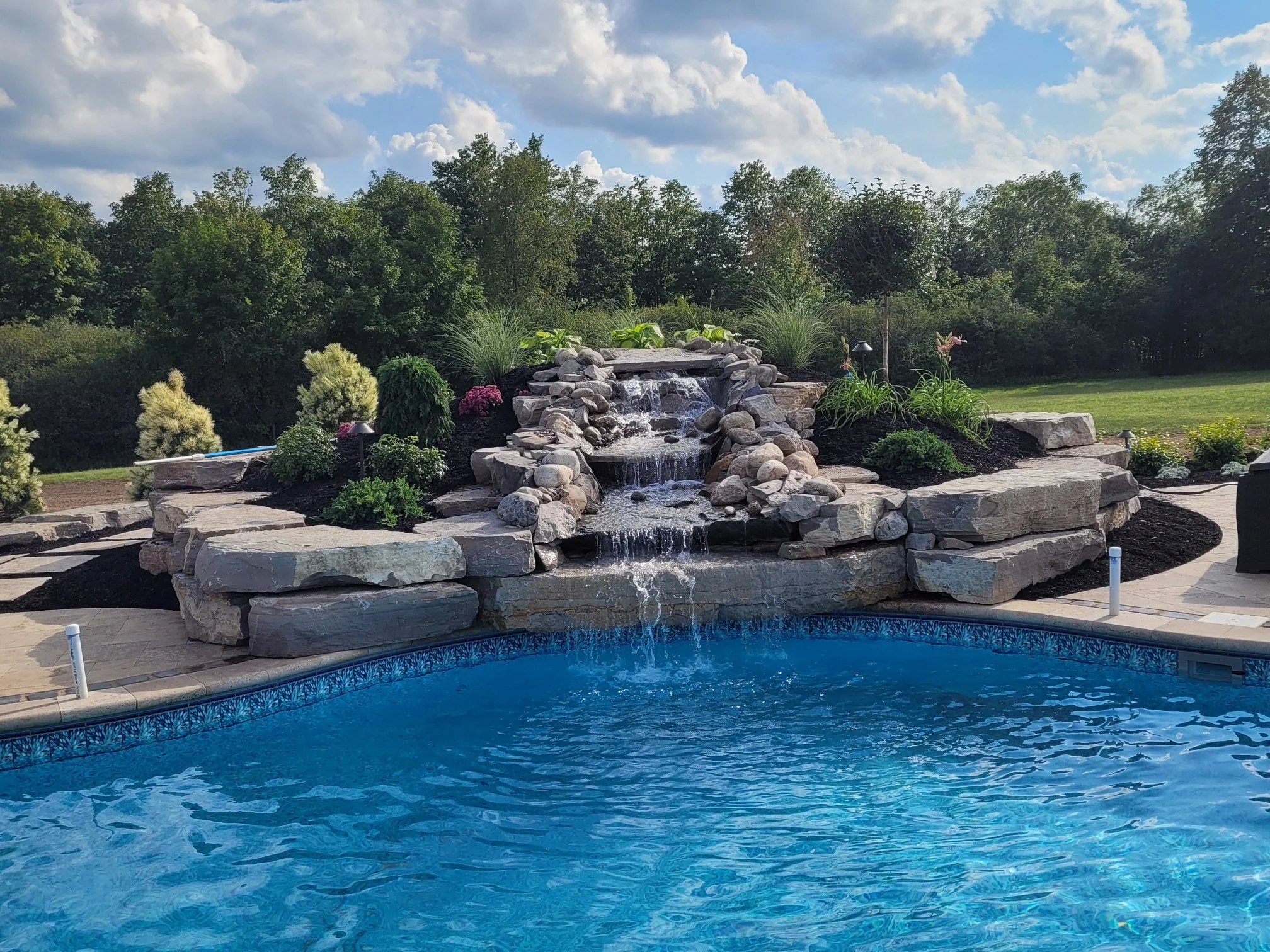 A backyard with a blue swimming pool and a stone waterfall feature surrounded by landscaped plants and trees under a partly cloudy sky.