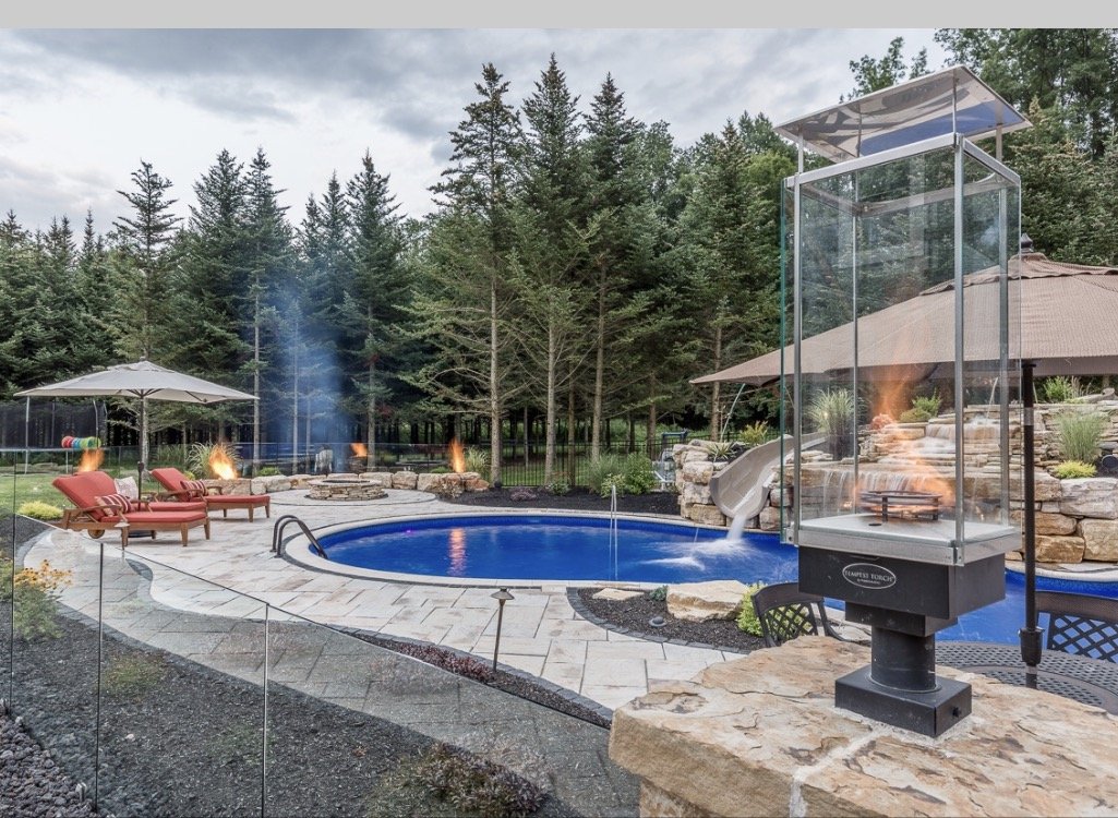 An outdoor backyard pool area surrounded by trees, with lounge chairs, umbrellas, fire pits, a waterslide, and a glass fire pit.