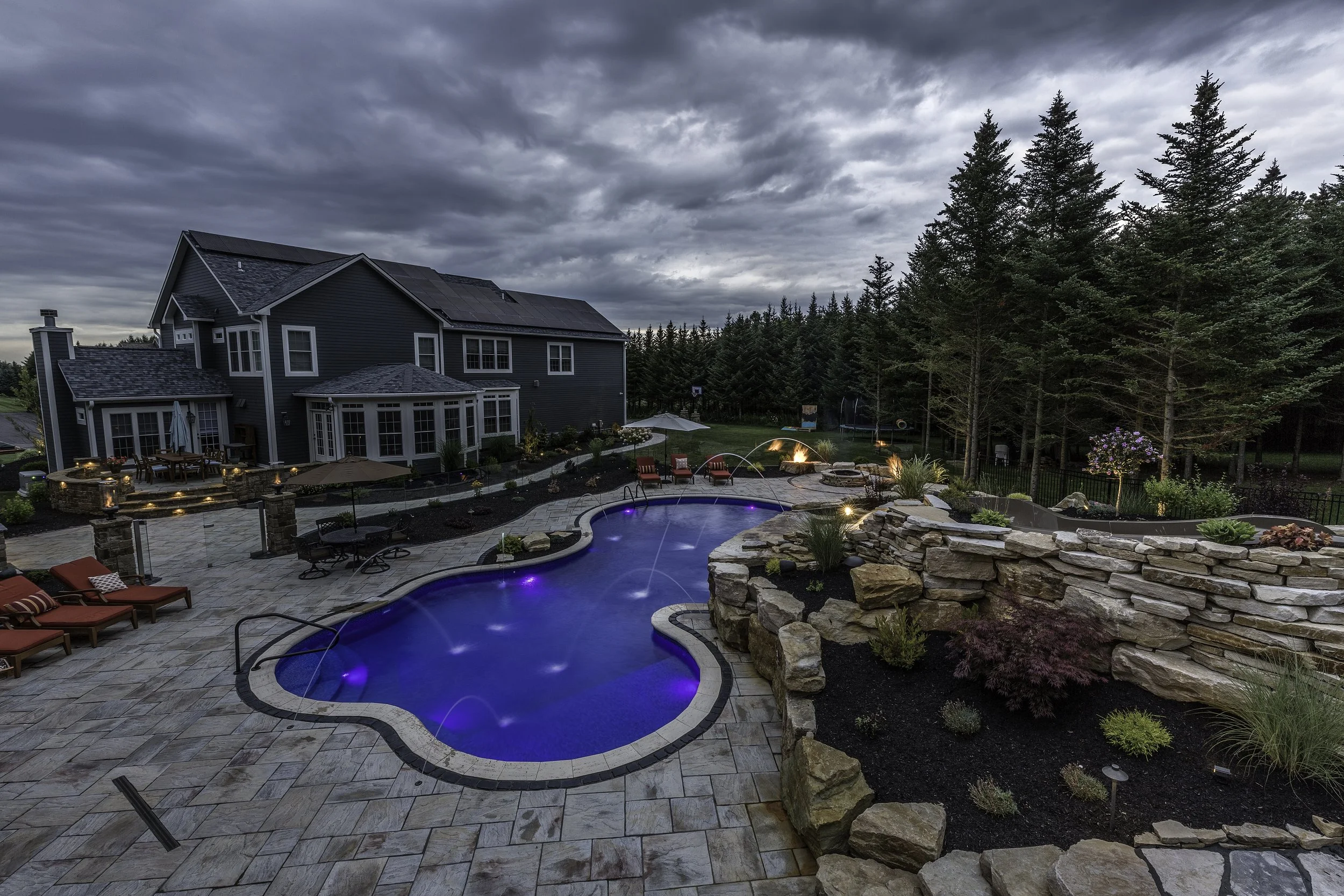 A backyard with a pool, lounge chairs, and a patio area, surrounded by a garden with trees and a large house, under a cloudy sky.