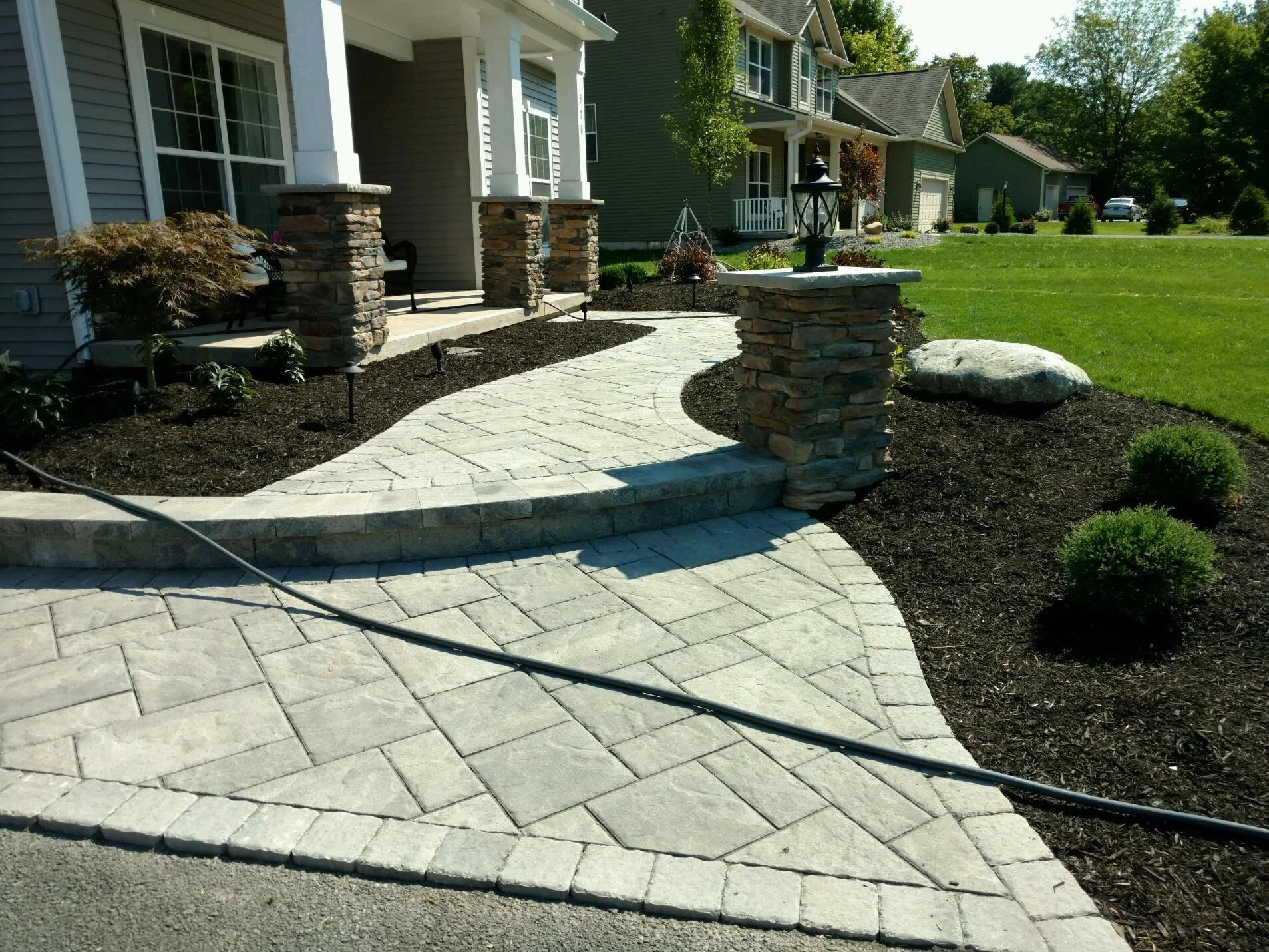Newly paved stone pathway leading to the front porch of a house, flanked by stone columns and landscaped with bushes and plants.