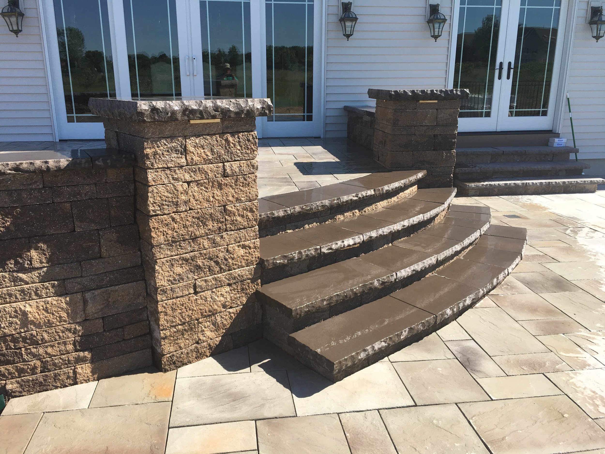 Exterior view of a house with a stone staircase leading to a patio. The patio has light-colored paving stones and a glass sliding door with wall lanterns on either side.
