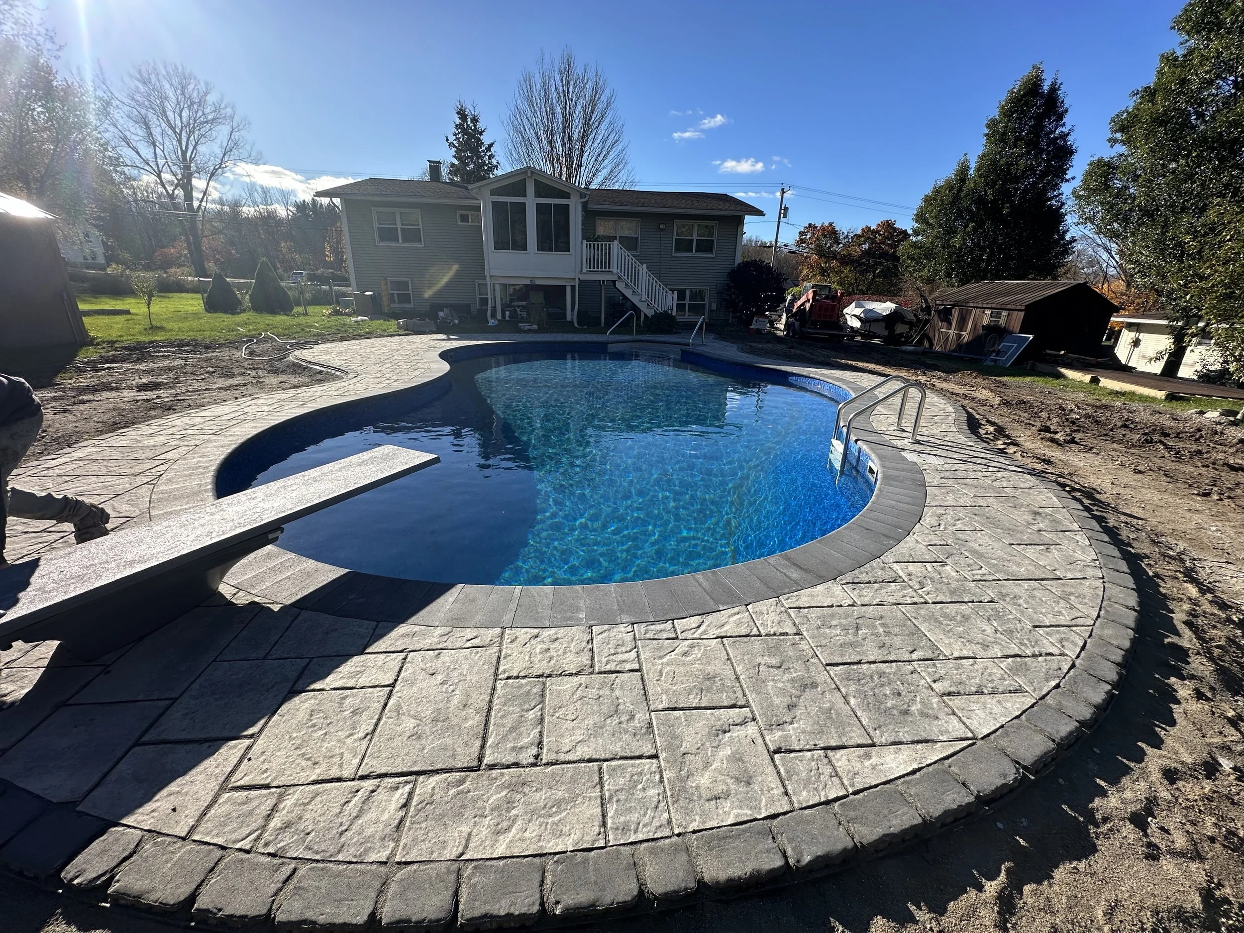In-ground swimming pool with a concrete deck, metal ladder, and a small bench, surrounded by grass and trees, with a house and other structures in the background on a sunny day.