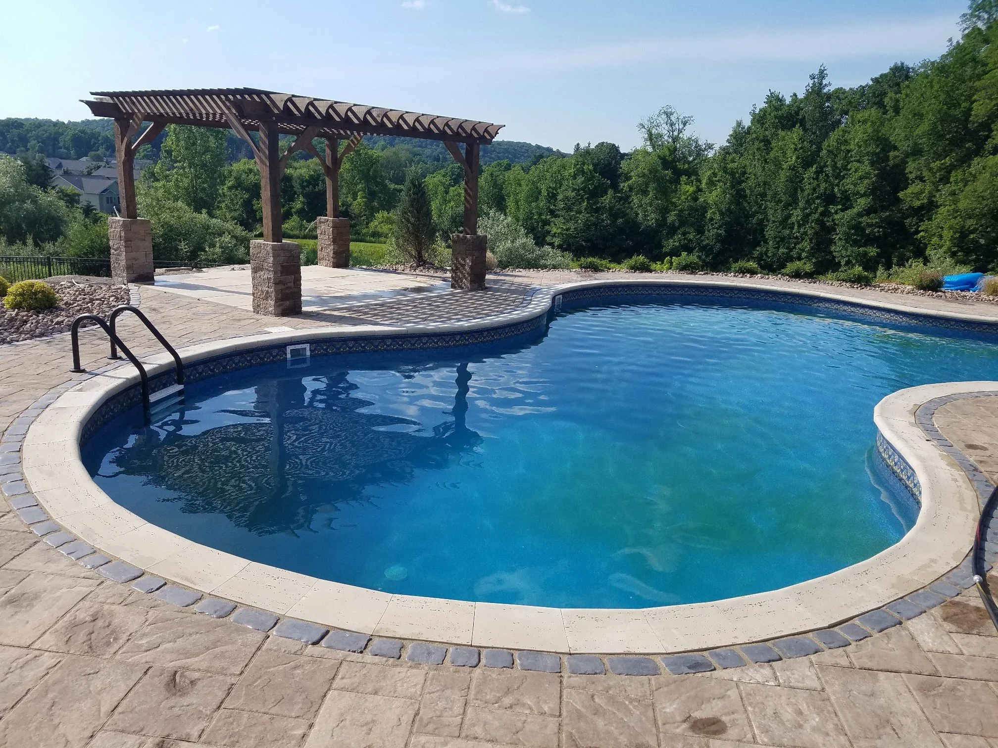 An outdoor swimming pool with a curved shape, surrounded by a stone patio. There is a wooden pergola with stone pillars providing partial shade. The background features lush green trees under a clear blue sky.