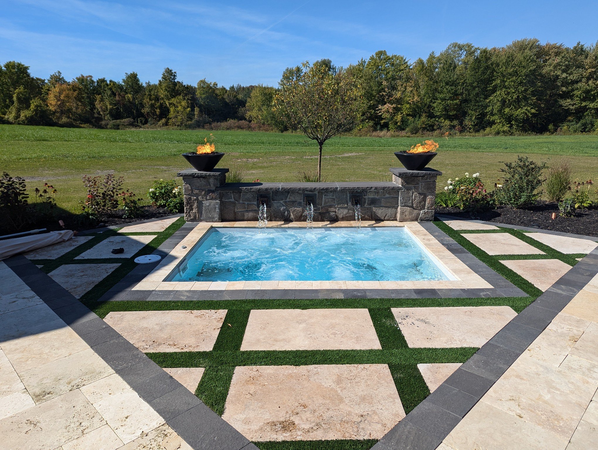 A backyard with a hot tub, surrounded by a stone patio with grass inlay, and decorative planters with fire on tall stone pedestals, set against a backdrop of open grassy area and trees under a blue sky.