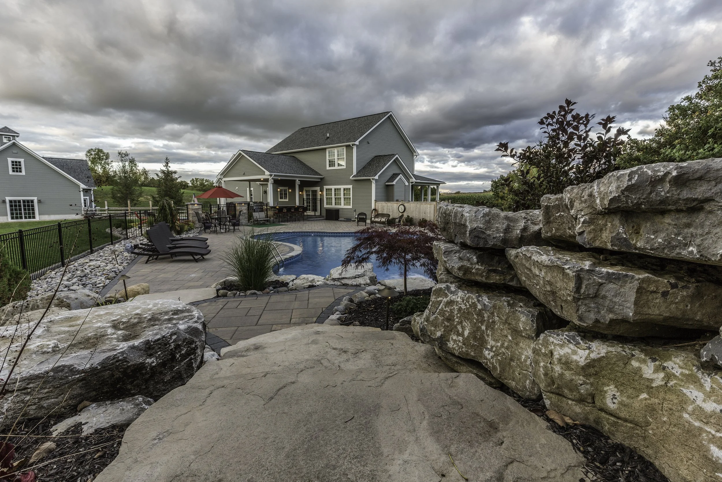 A backyard with a swimming pool, lounge chairs, a table with a red umbrella, and a two-story house under a cloudy sky.