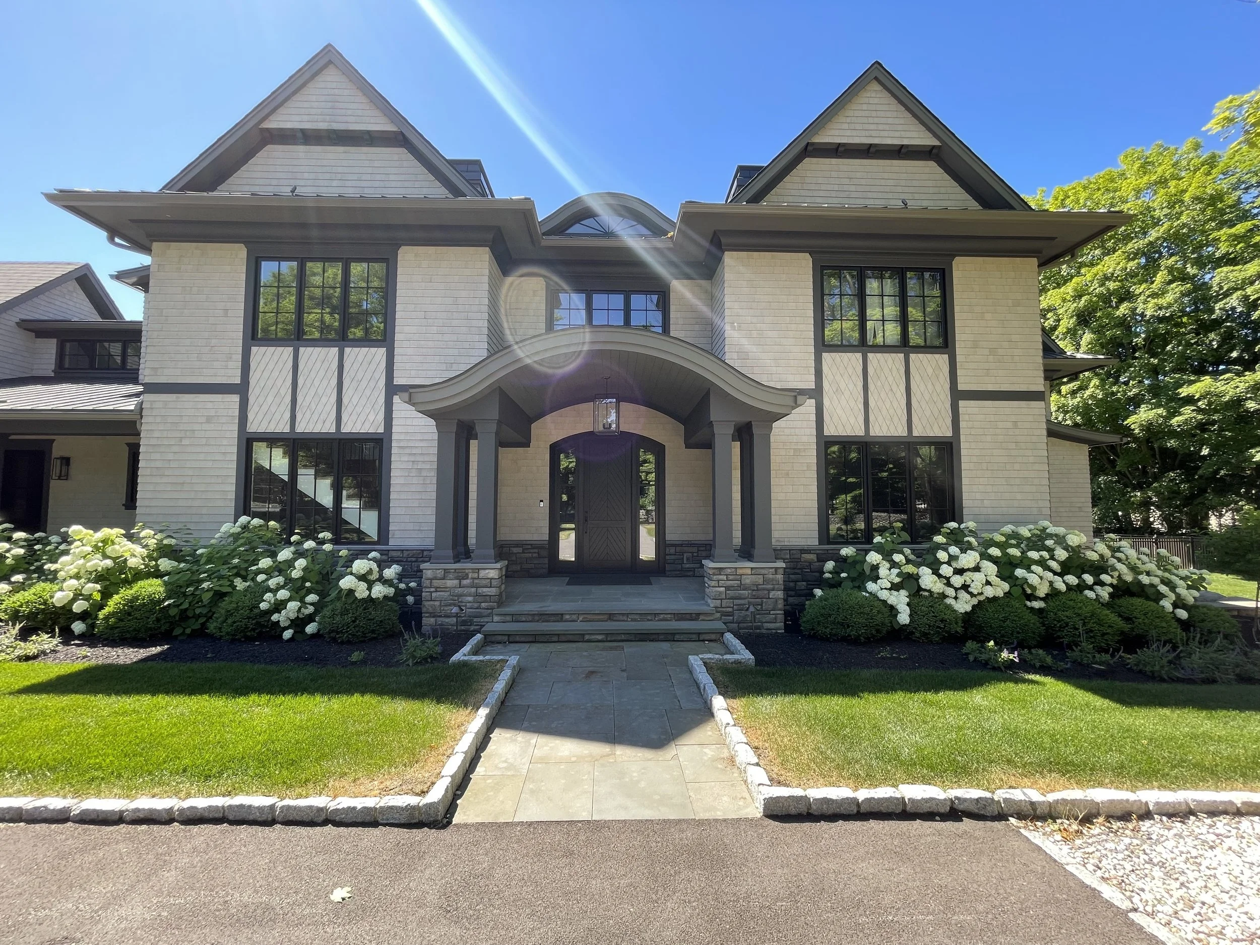 Front view of a large two-story house with beige siding and black trim, with a manicured lawn and white hydrangea flowers in the garden beds. Sunlight creates a lens flare effect.
