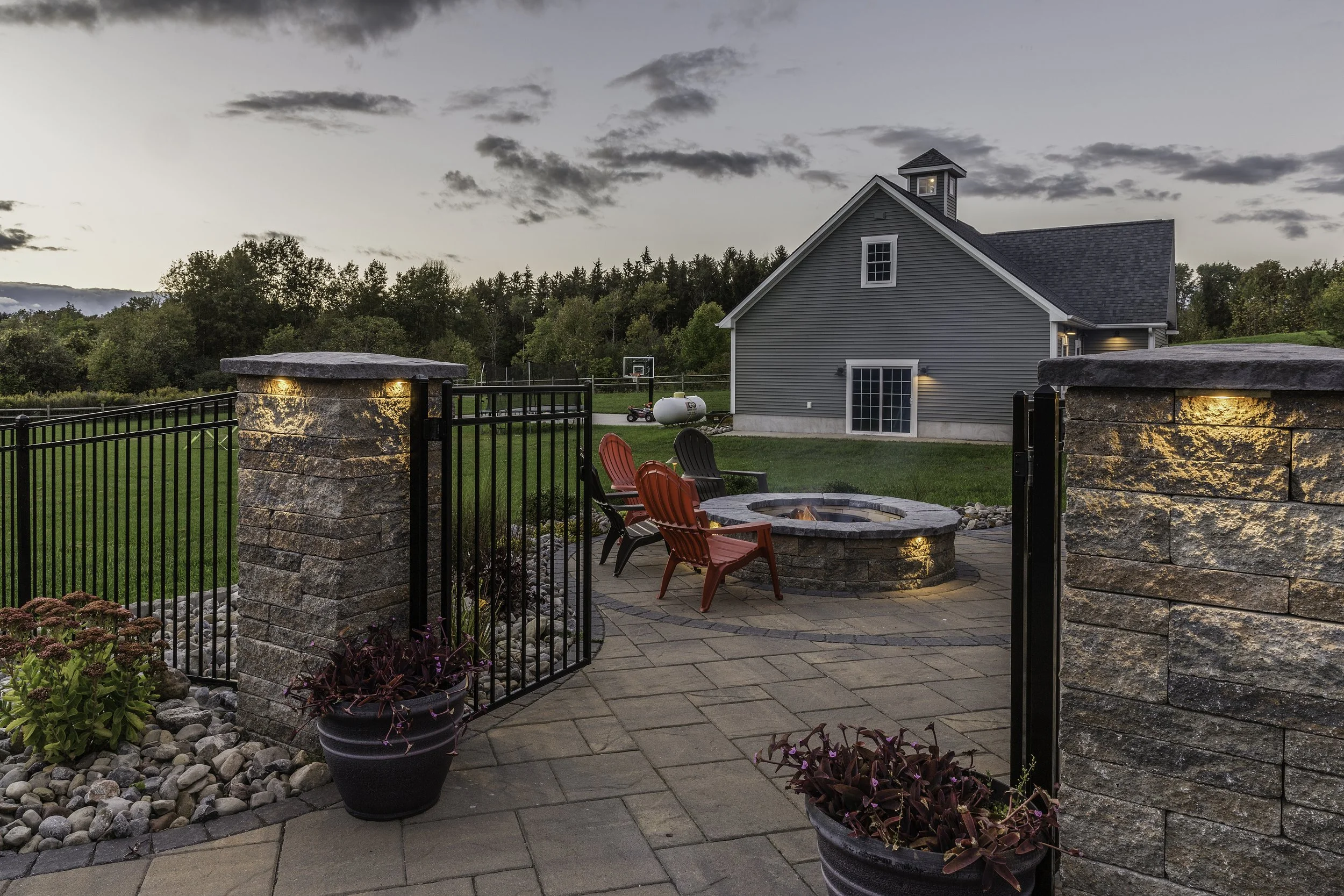 A backyard patio with a fire pit, red and black Adirondack chairs, potted plants, and a view of a gray house, green field, and trees in the distance during dusk.