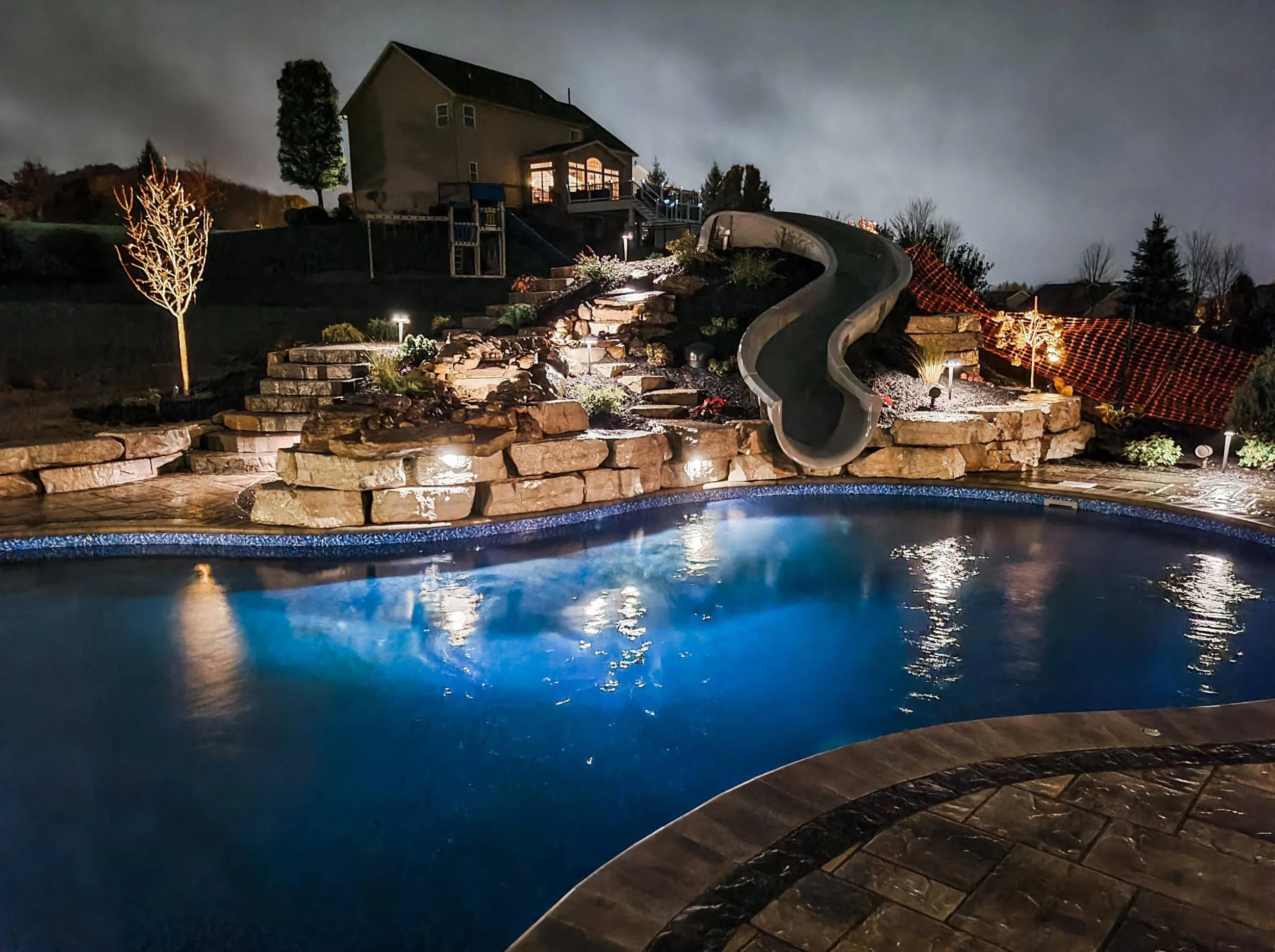 Nighttime view of a backyard with a swimming pool, waterfall, and a water slide, illuminated by outdoor lights, with house and trees in the background.