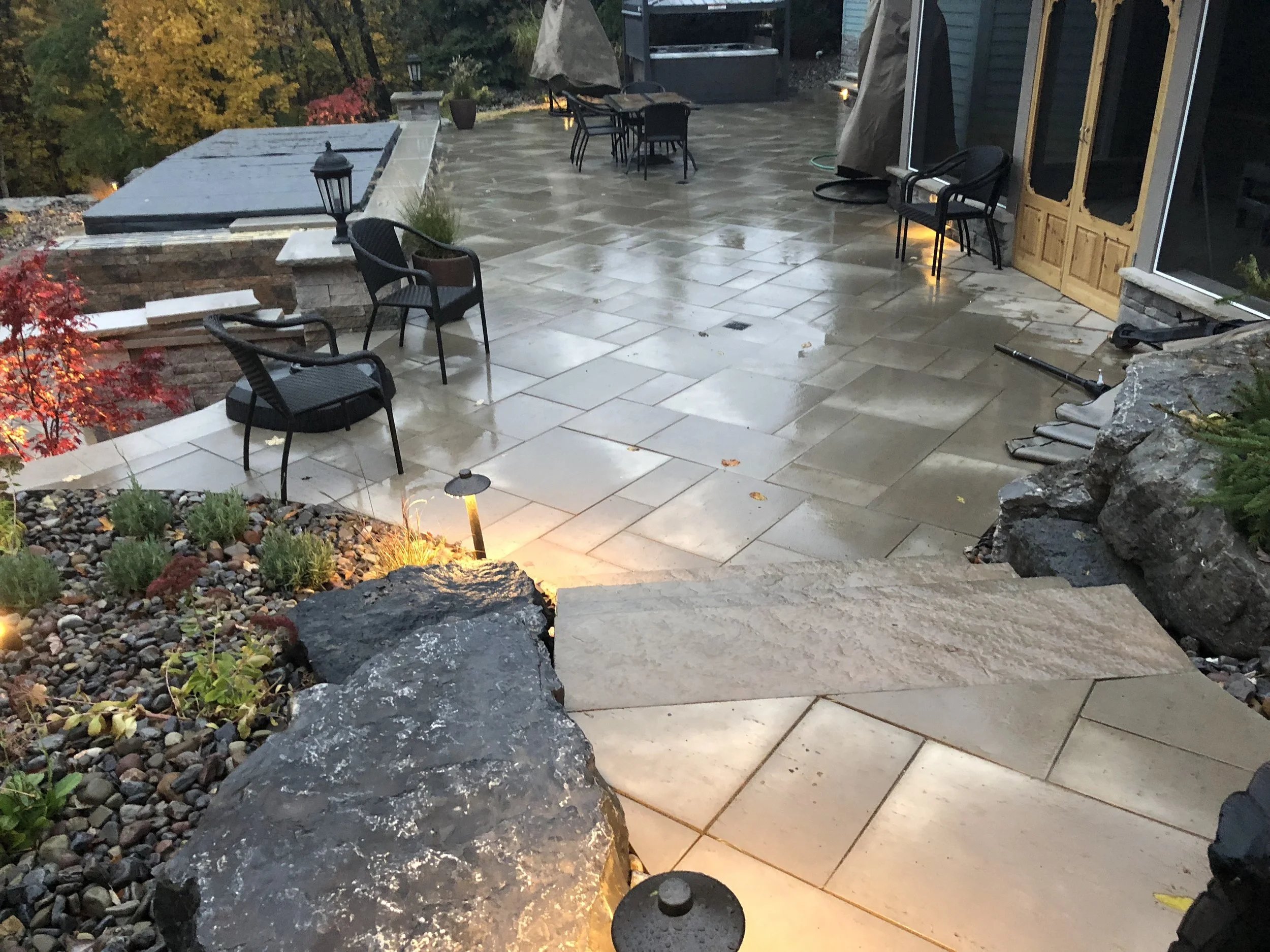 Wet outdoor patio with black metal chairs, stone pavers, smaller rocks with plants, and star-shaped landscape lights