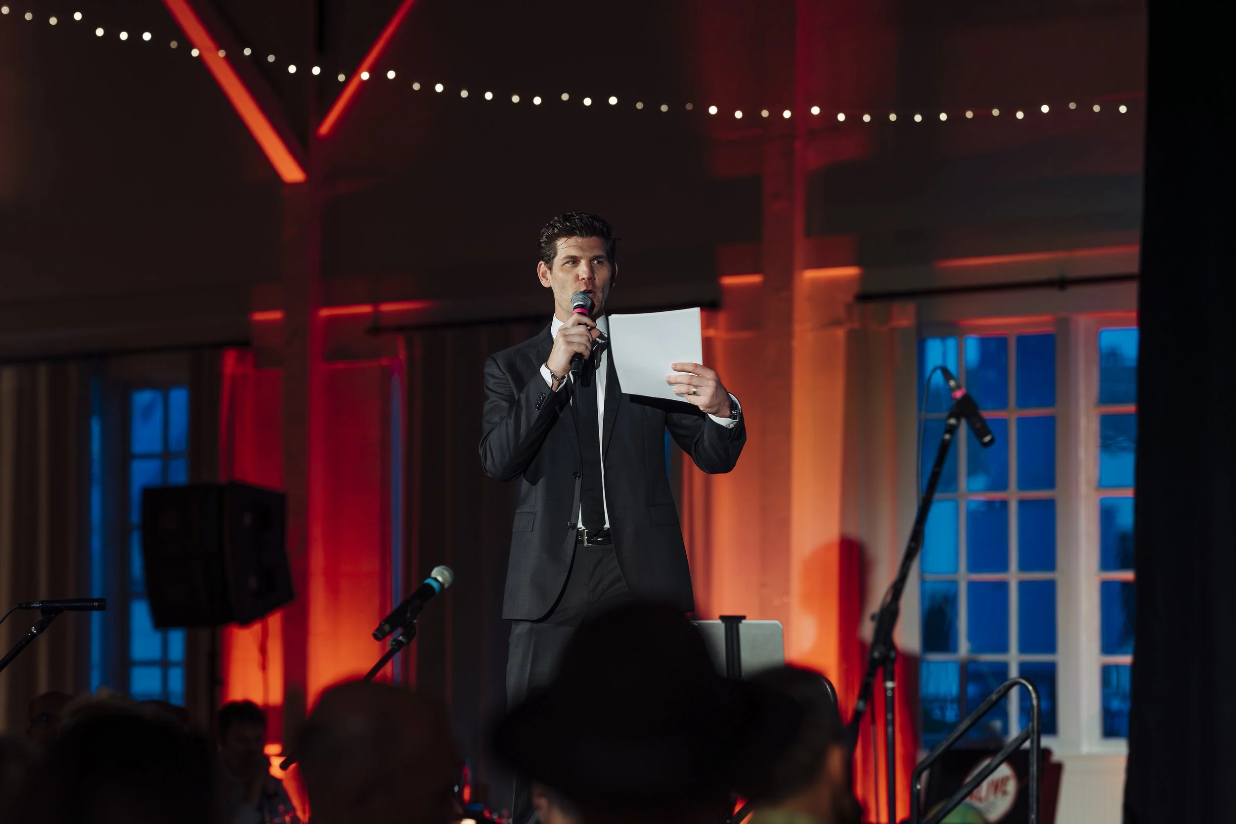 A man in a black suit and white shirt holding a microphone and a sheet of paper, speaking at an event with warm lighting and a decorated background.