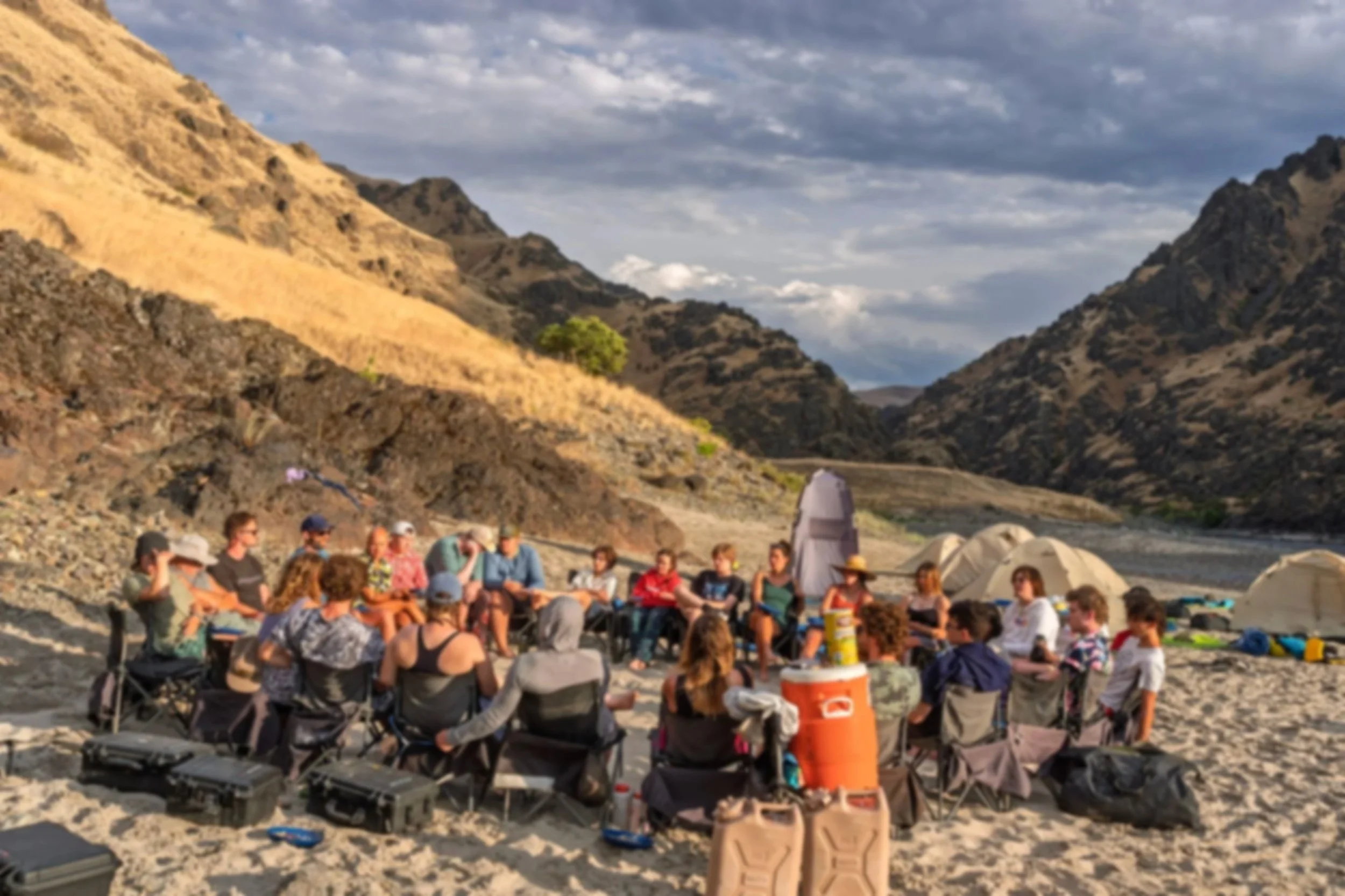 Group of people sitting on chairs and logs near rocky terrain, with tents and backpacks nearby, in a mountainous area with cloudy sky.