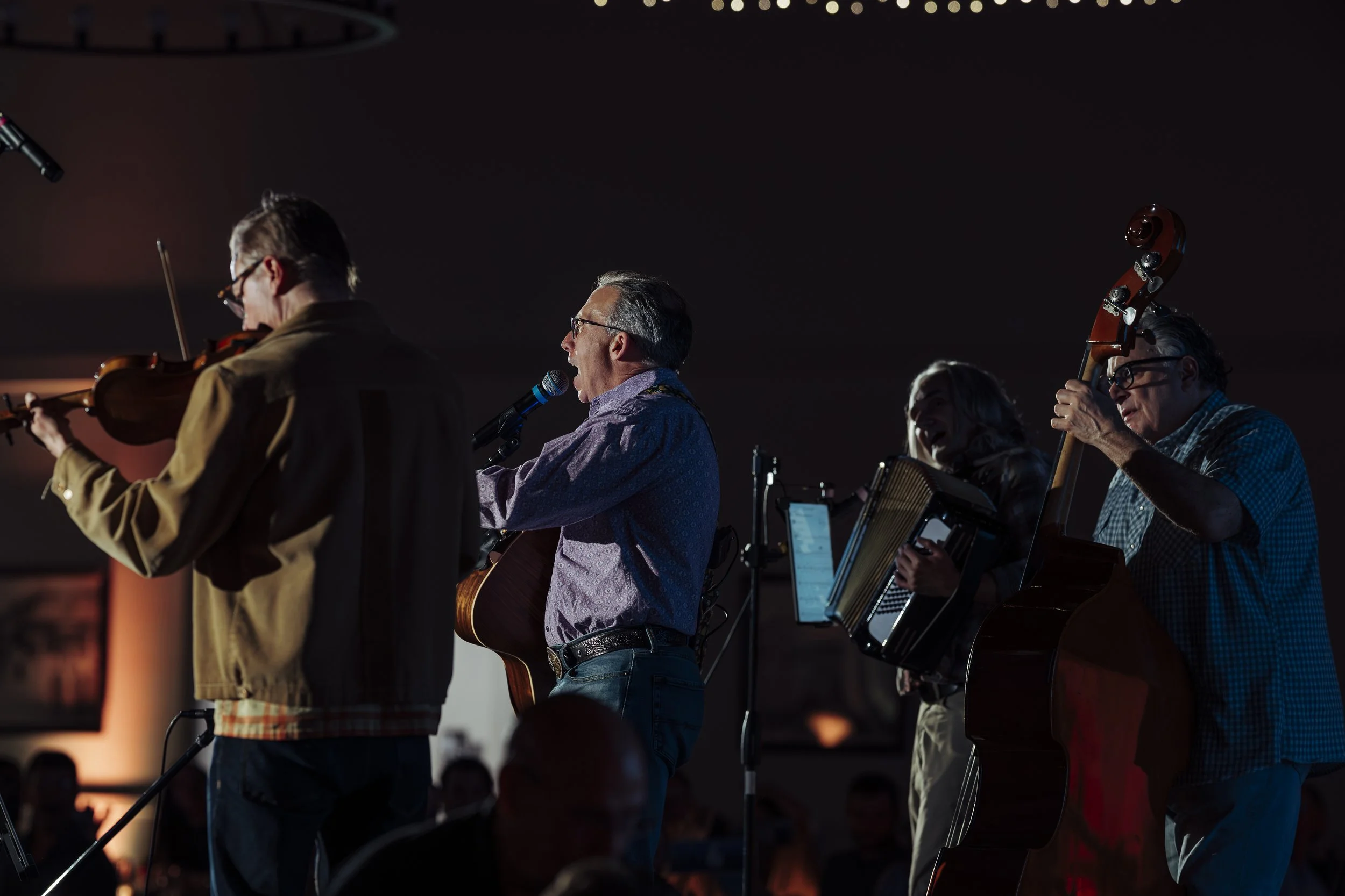 A group of musicians performing on stage, including a violinist, a singer with a microphone, and a guitarist, with a dark background.