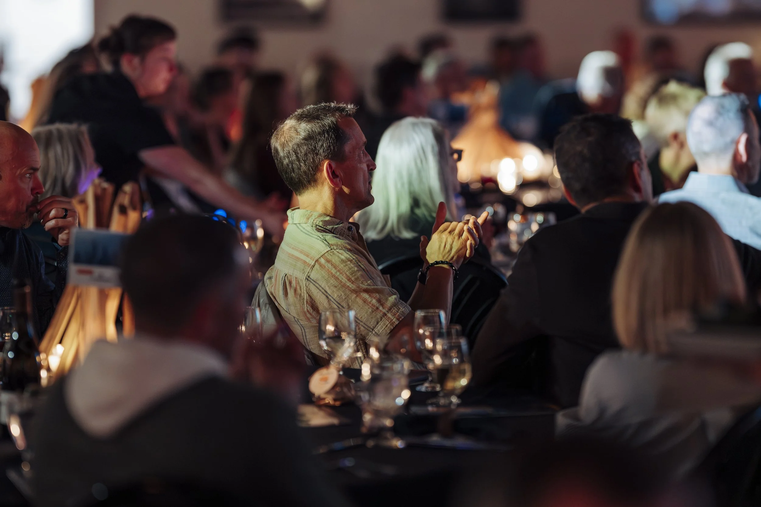 People attending the Variety Show, seated at tables, watching the mission video, in a dimly lit room.