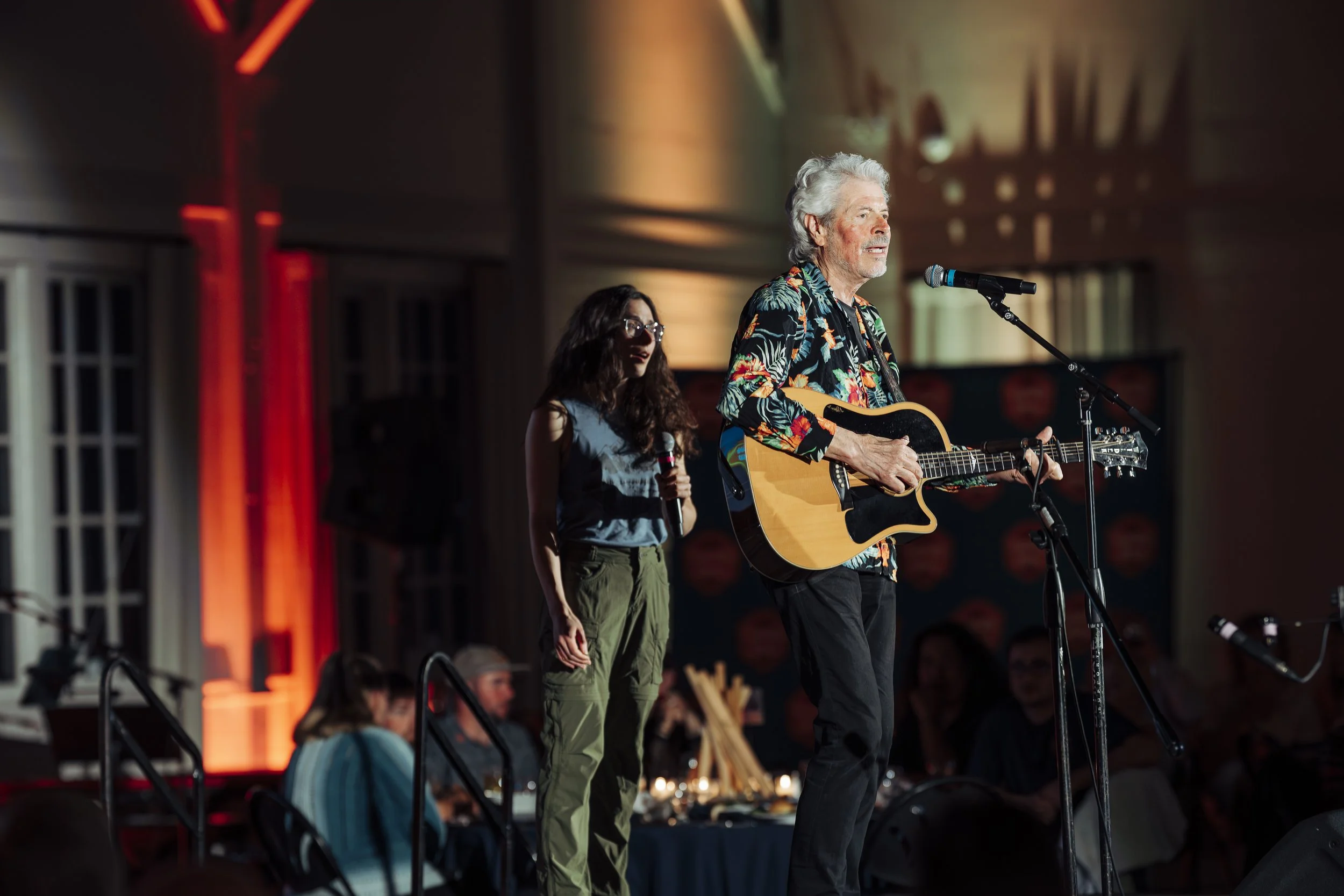 A performer playing an acoustic guitar on stage, accompanied by a second performer holding a microphone. The stage is lit with warm, colorful lighting, and there are audience members visible in the background.