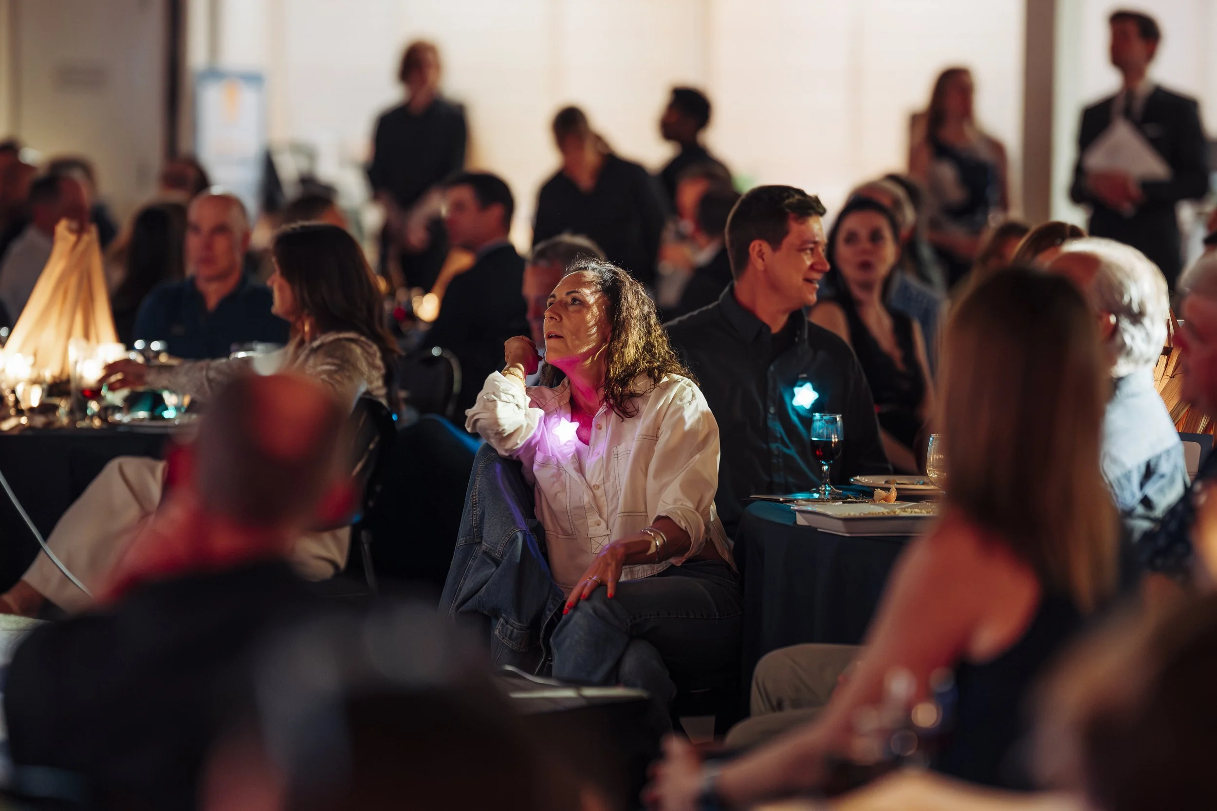 People gathered in a dimly lit banquet hall, some seated at tables with plates and drinks, others standing and chatting, with a woman with curly hair and a white shirt in focus, smiling and looking upward.