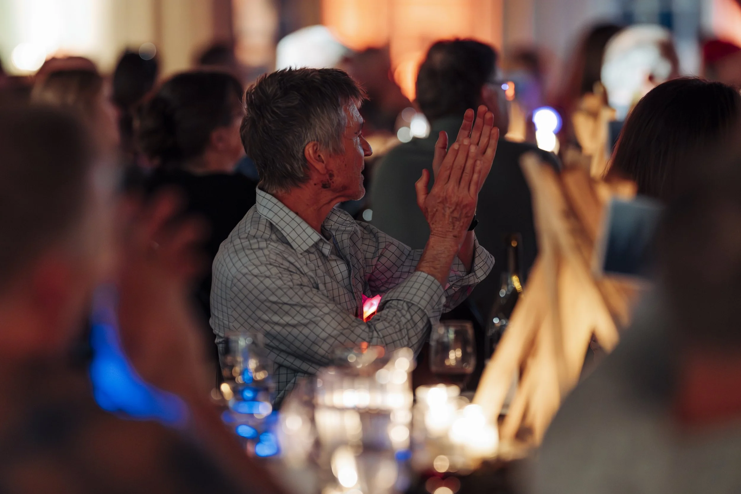 An person sitting at a table, prayer hands together, in a dimly lit setting with other people around, possibly at the Variety Show.