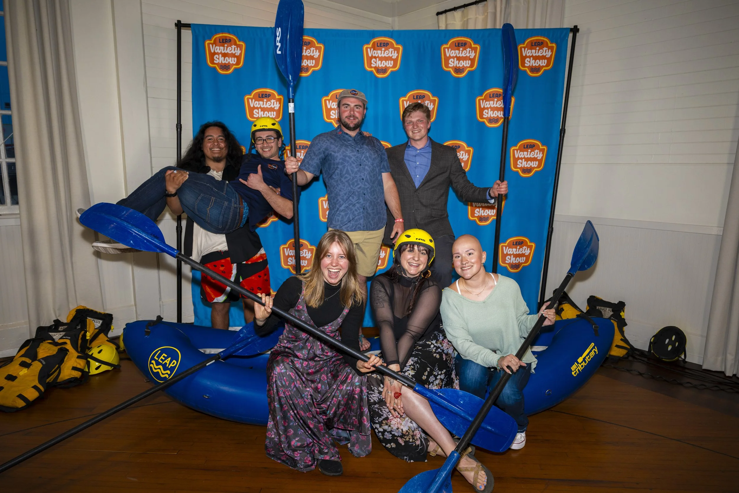 A group of seven people posing inside a room, with a blue background featuring the text 'LEAP Variety Show' repeatedly. Some are holding paddles, and others are wearing yellow helmets and colorful clothing. There is a blue inflatable boat at their fe