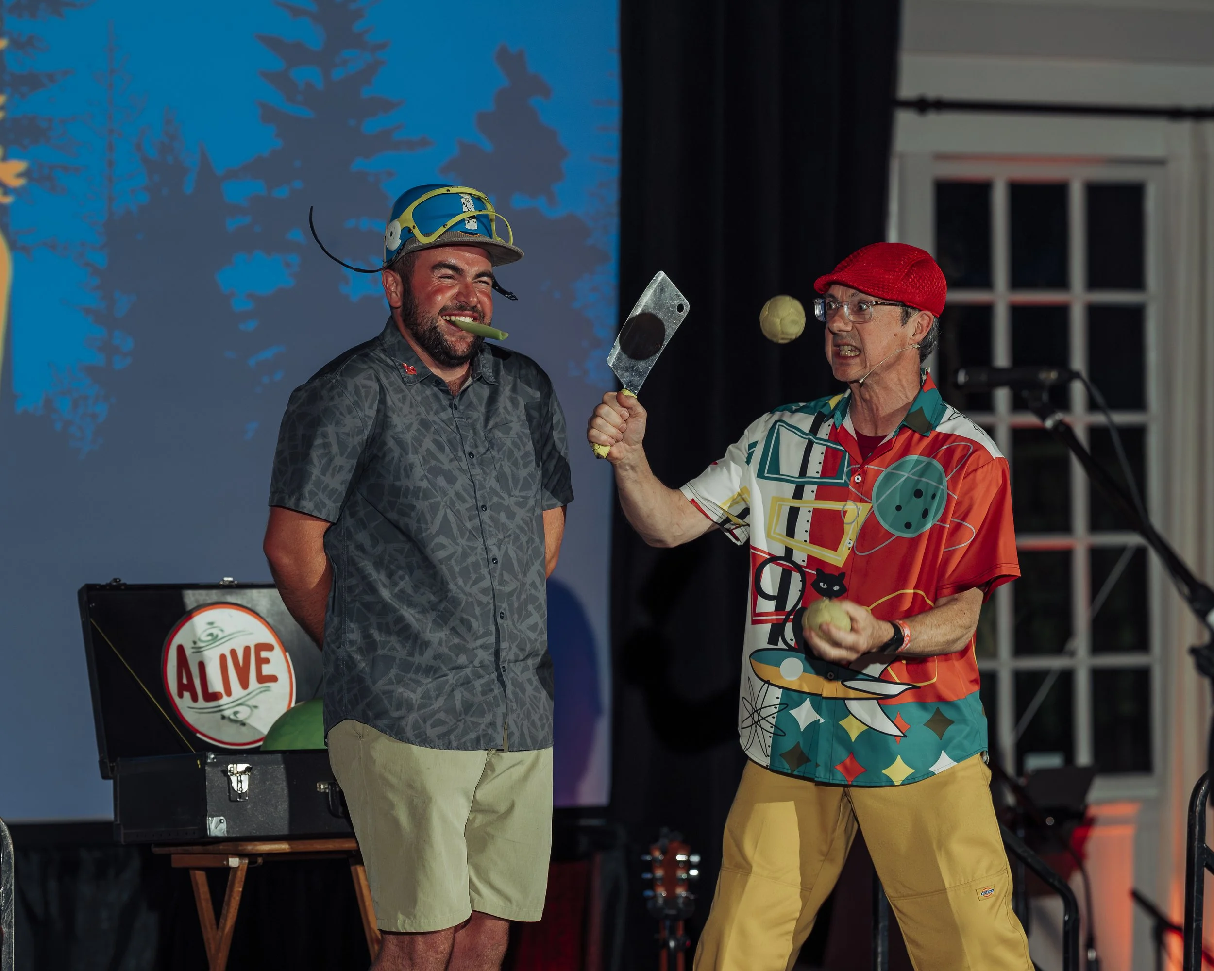 Two people stand indoors on a stage: one dressed as a clown and the other wearing a helmet with a whistle. The person dressed as a clown is holding a tennis ball and appears to be in a playful confrontation with the person wearing the helmet.