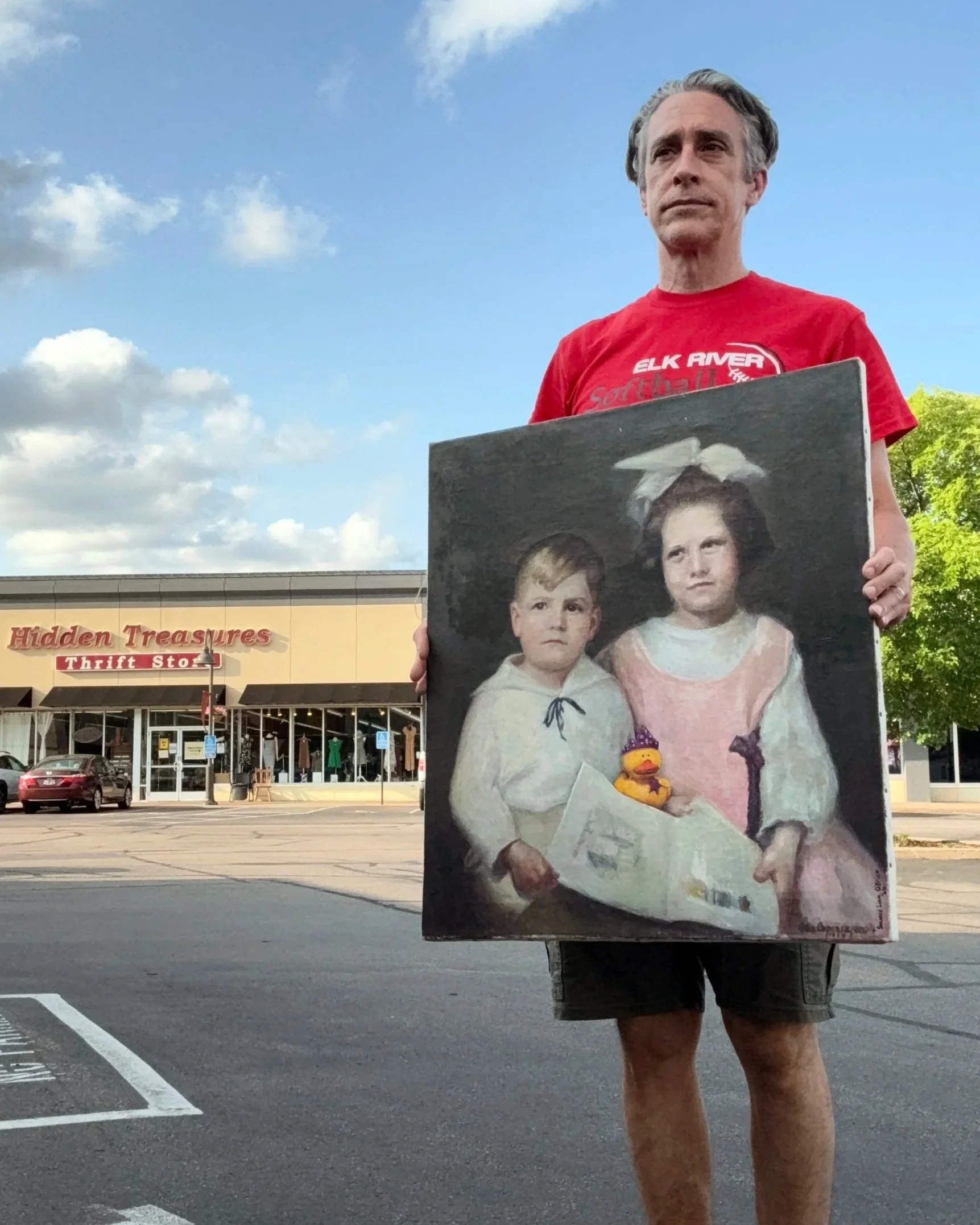 Man wearing a red shirt holding a portrait painting with a rubber duck in the parking lot of a thrift store.