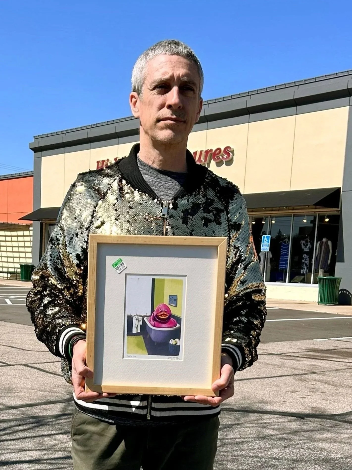 Man wearing a glitter jacket holding a framed painting of a large pink rubber duck in a bathtub.