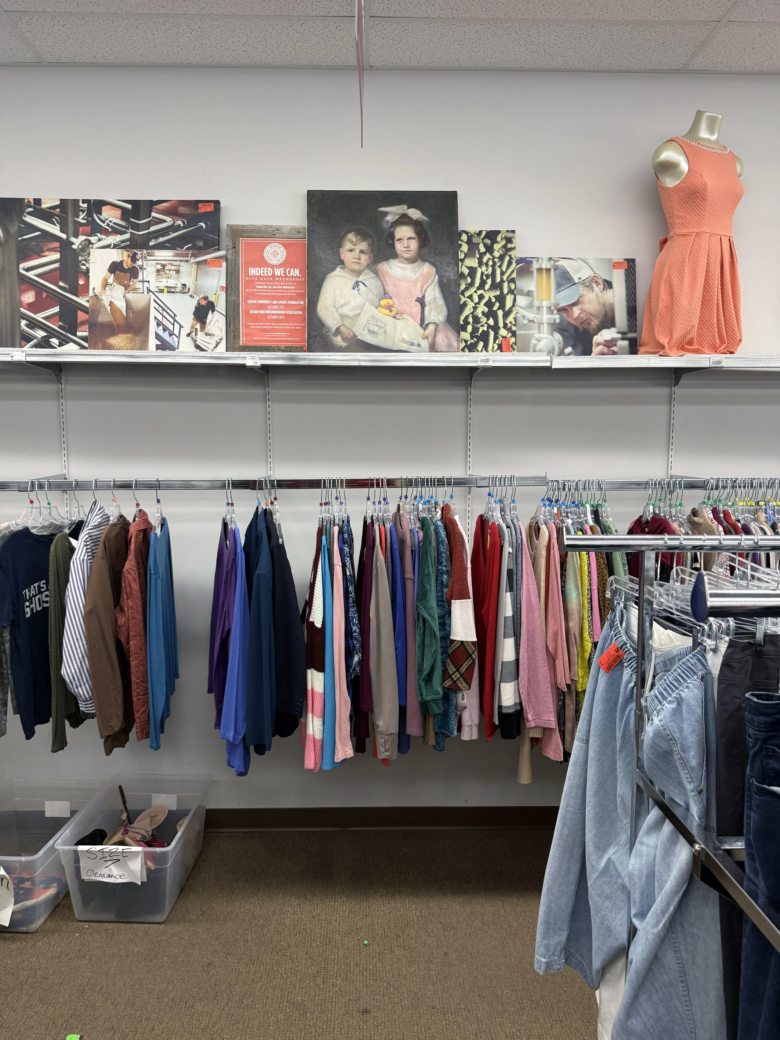Numerous paintings sitting on a store shelf above a hanger rack with shirts and sweaters.