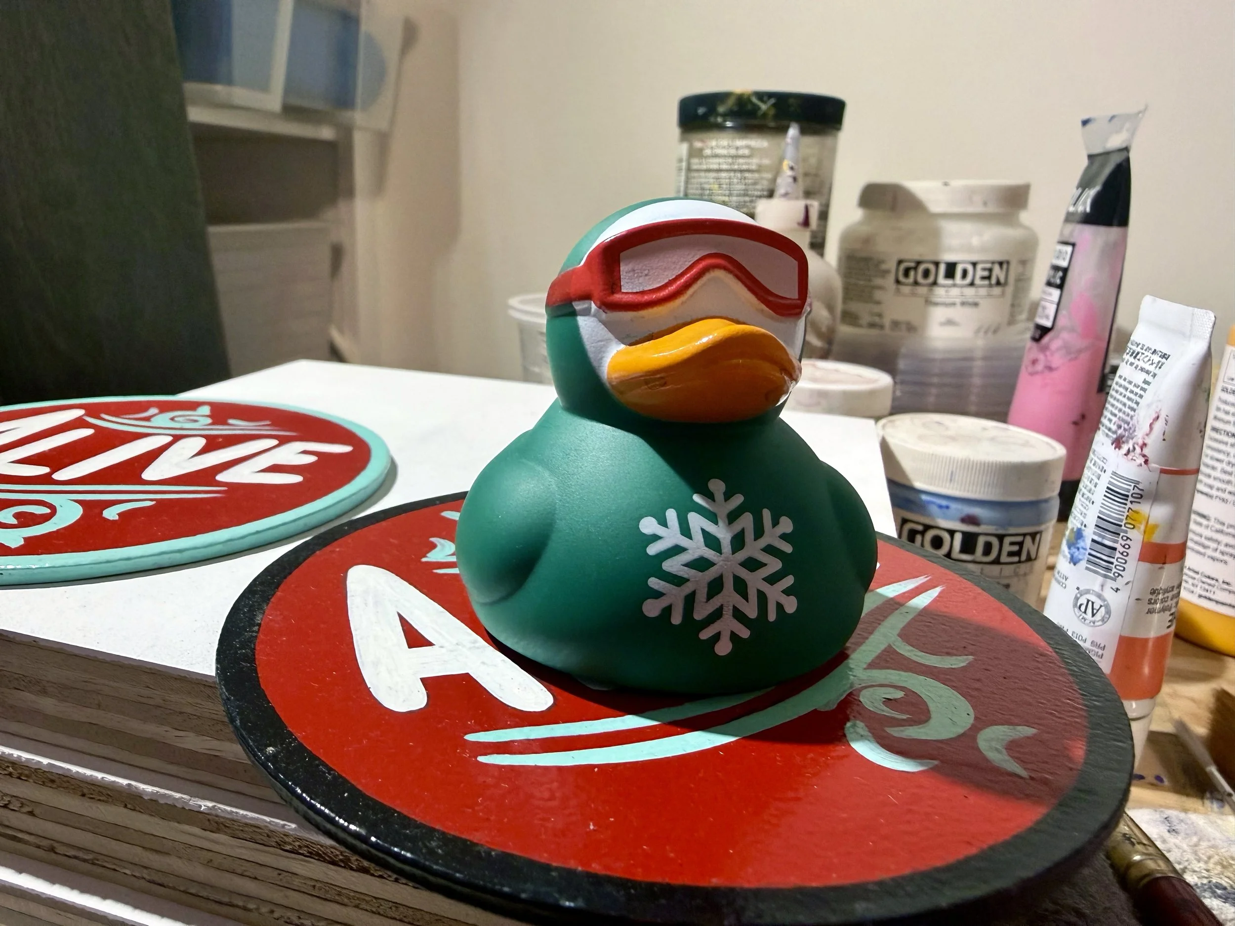 Rubber duck toy wearing a green wetsuit and goggles sitting on top of a red coaster in front of some paint.