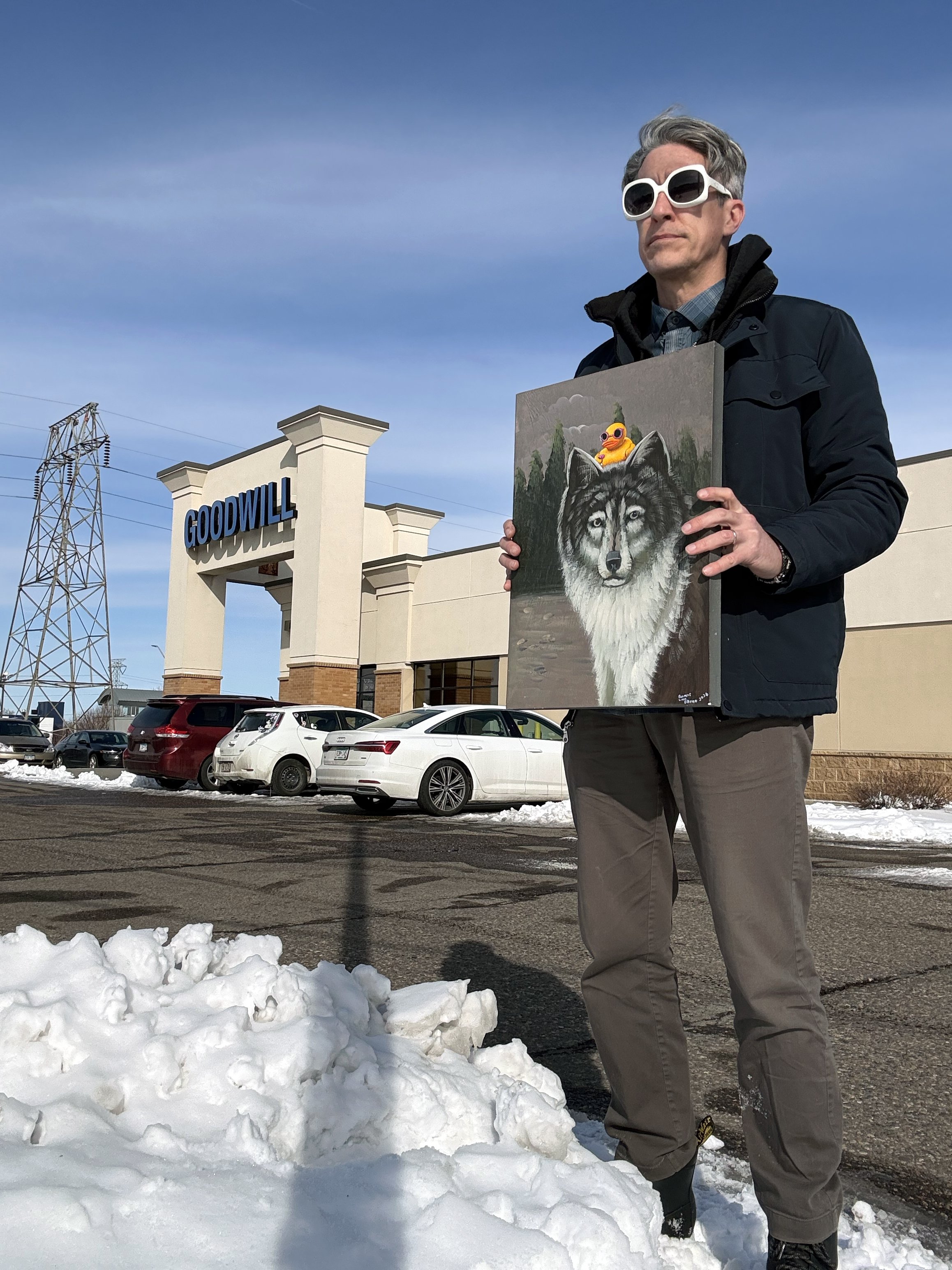Guy with sunglasses holding a painting of a wolf with a yellow rubber duck on its head in front of a Goodwill Thrift Store.