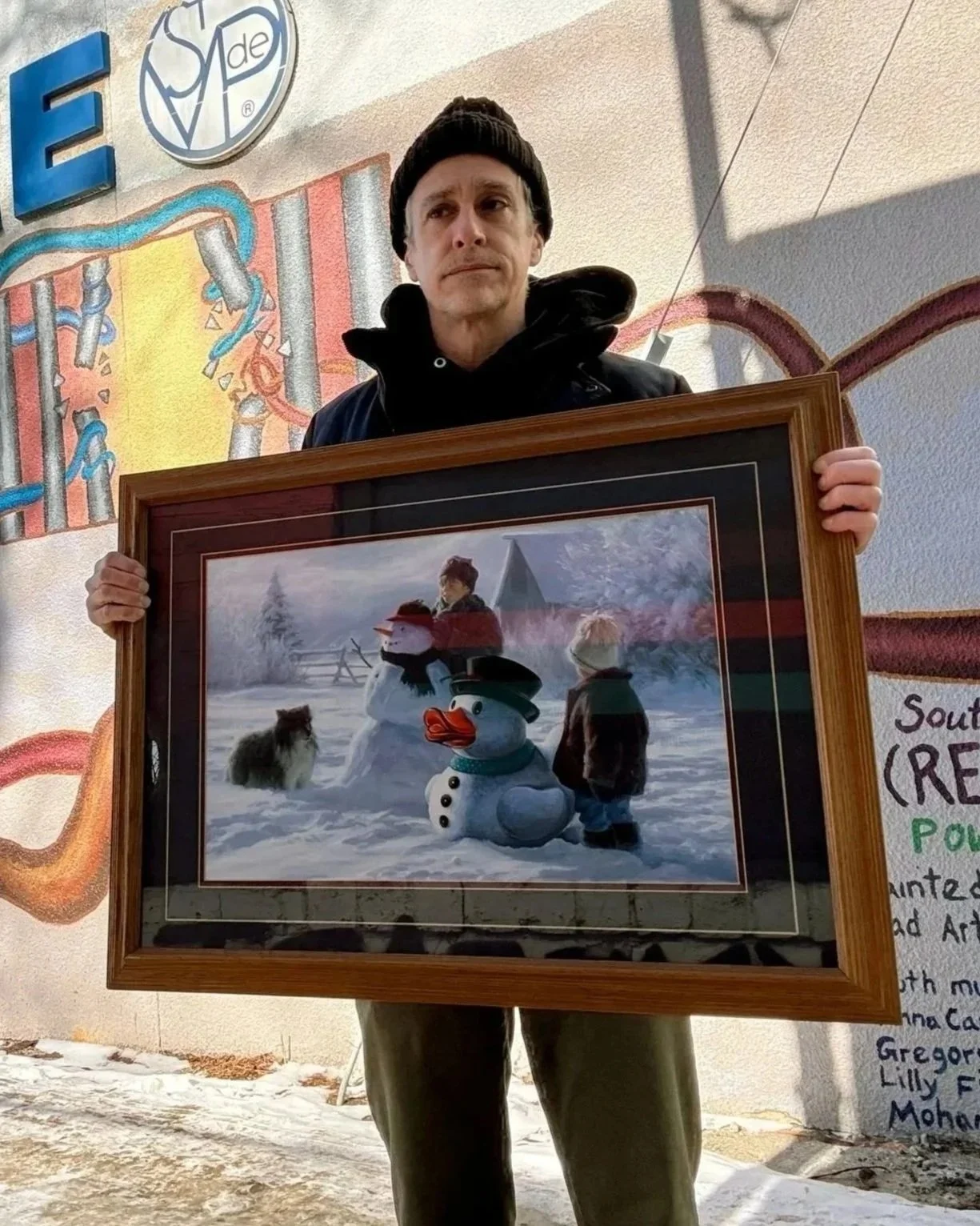 MAn holding a painting of a boy building a snowman next to a large white rubber duck
