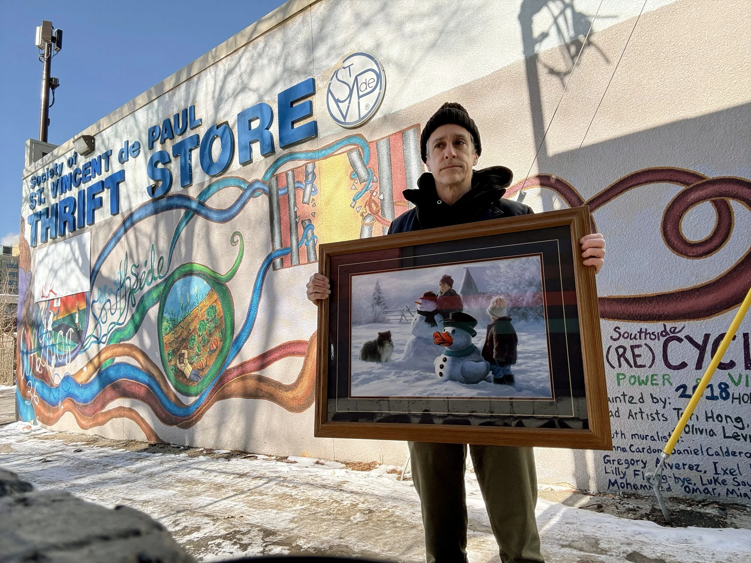 Man wearing a black sock hat holding a painting of a snowman and a white rubber duck in front of a thrift store.