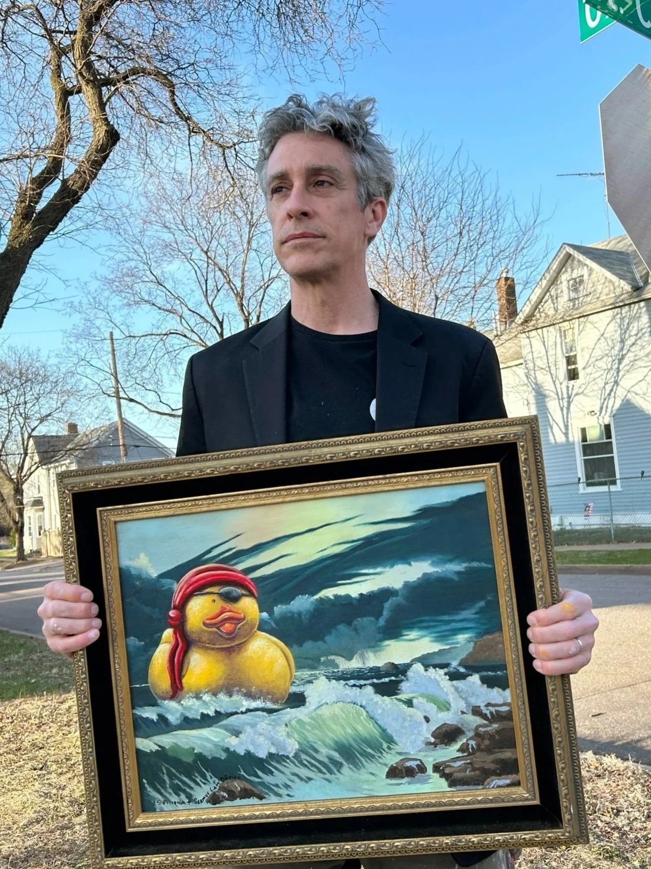 Man wearing a dinner jacket holding a painting of a rubber duck floating on the waves near a beach