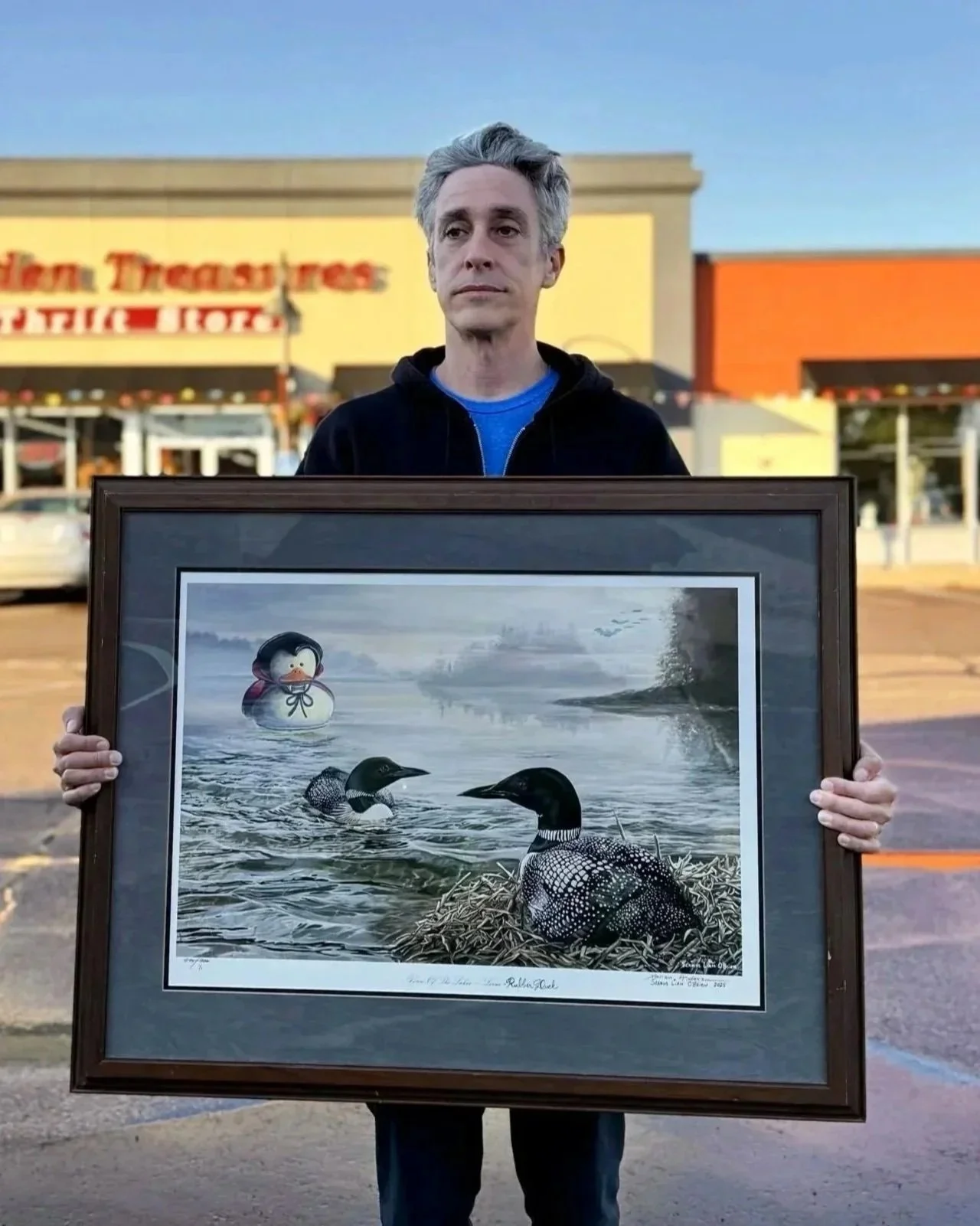 Man holding a framed painting of two loon birds floating in front of a large dracula rubber duck.