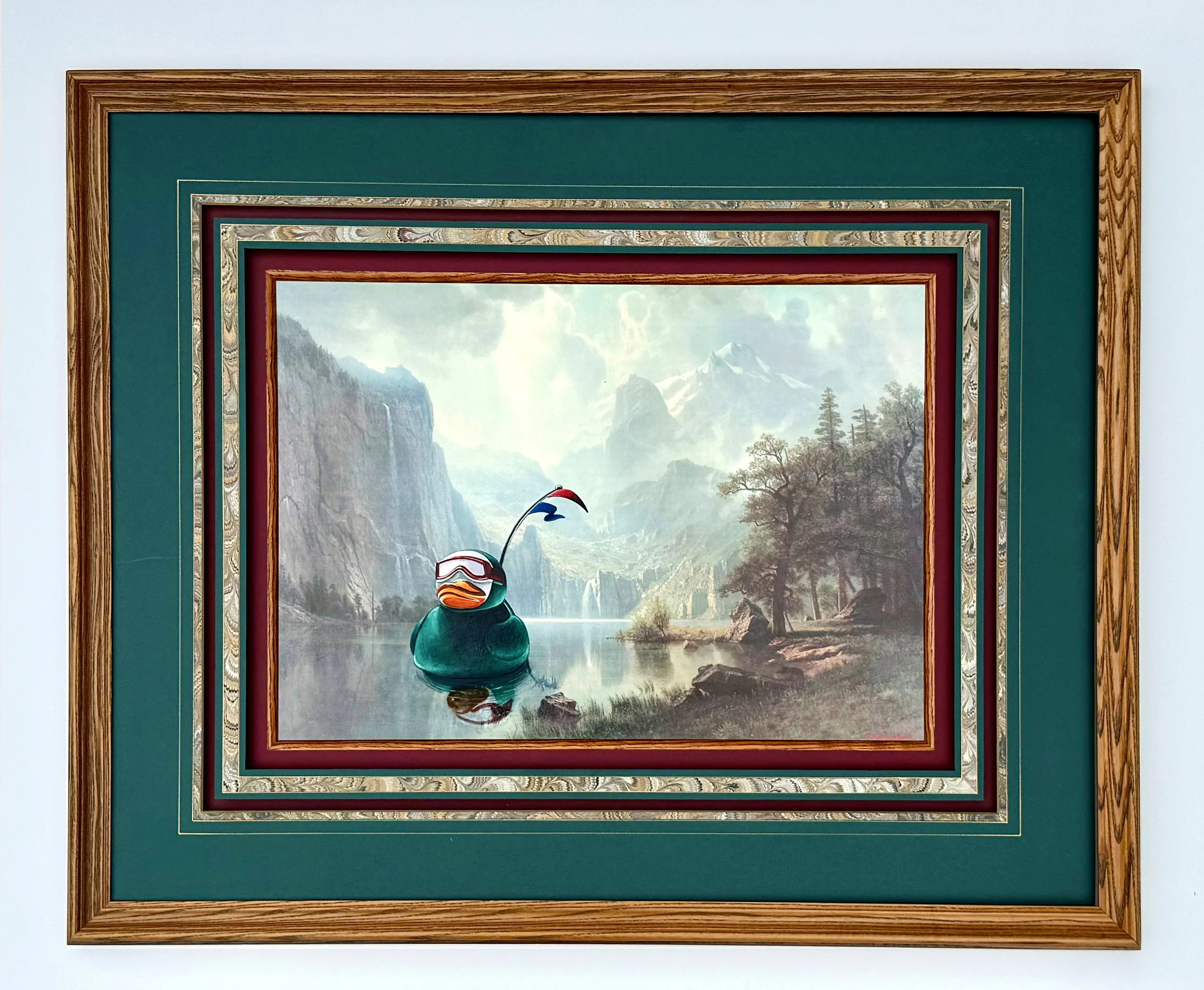 Framed painting with green matte board of a large rubber duck wearing a green wetsuit and goggles floating down a river in a monumental landscape.