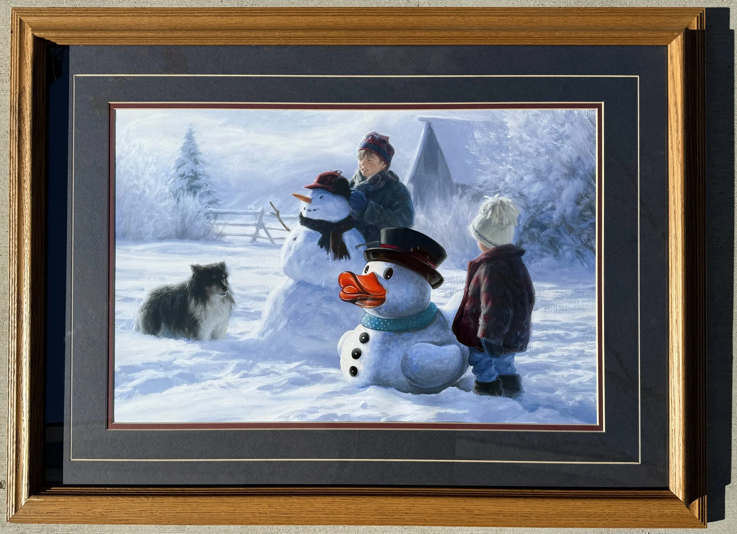 Framed painting of a boy building a snowman next to a large, white rubber duck while accompanied by a dog and another boy observing from the sides.