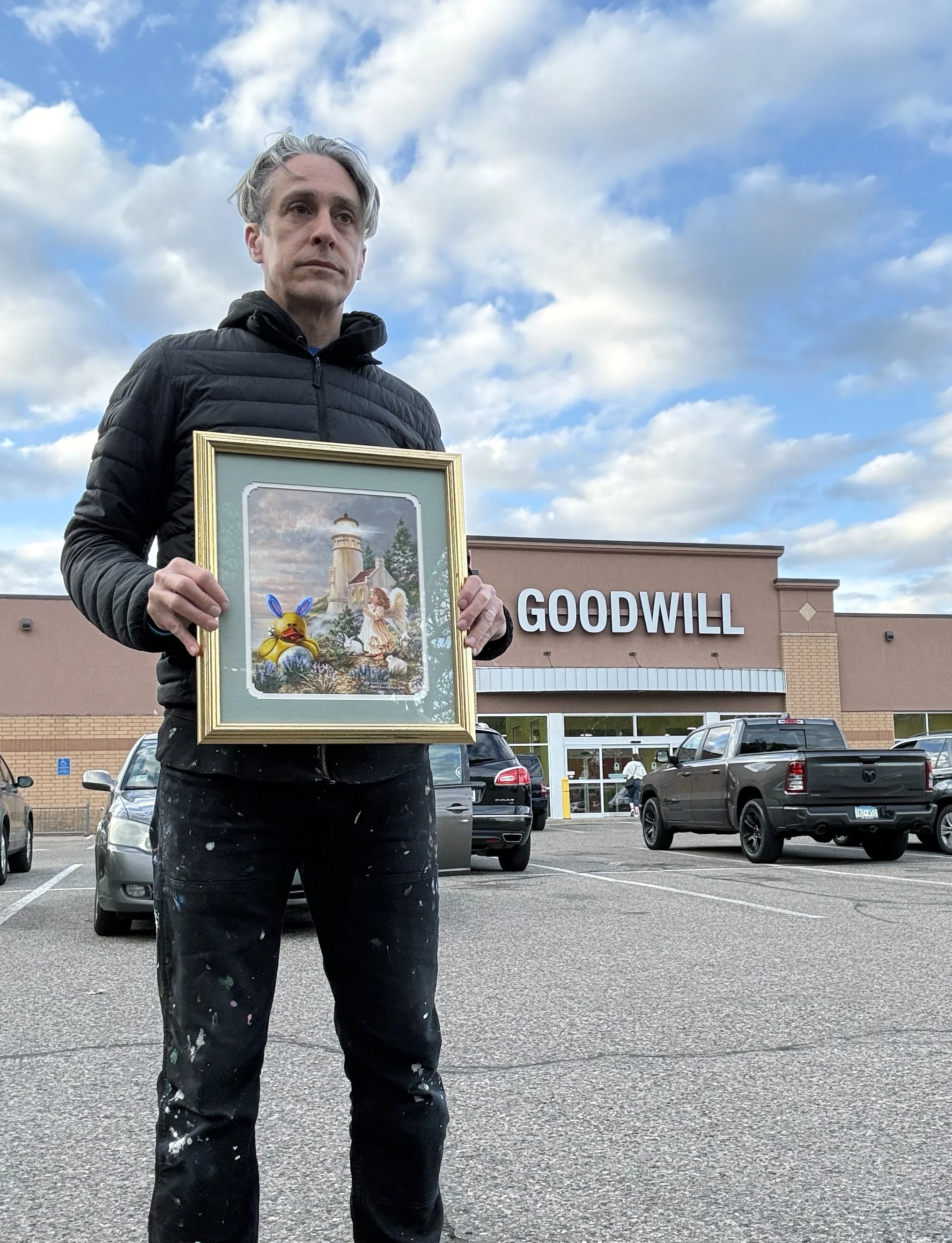 A man wearing a black jacket holding a colorful painting of a rubber duck in the parking lot of a Goodwill thrift store.