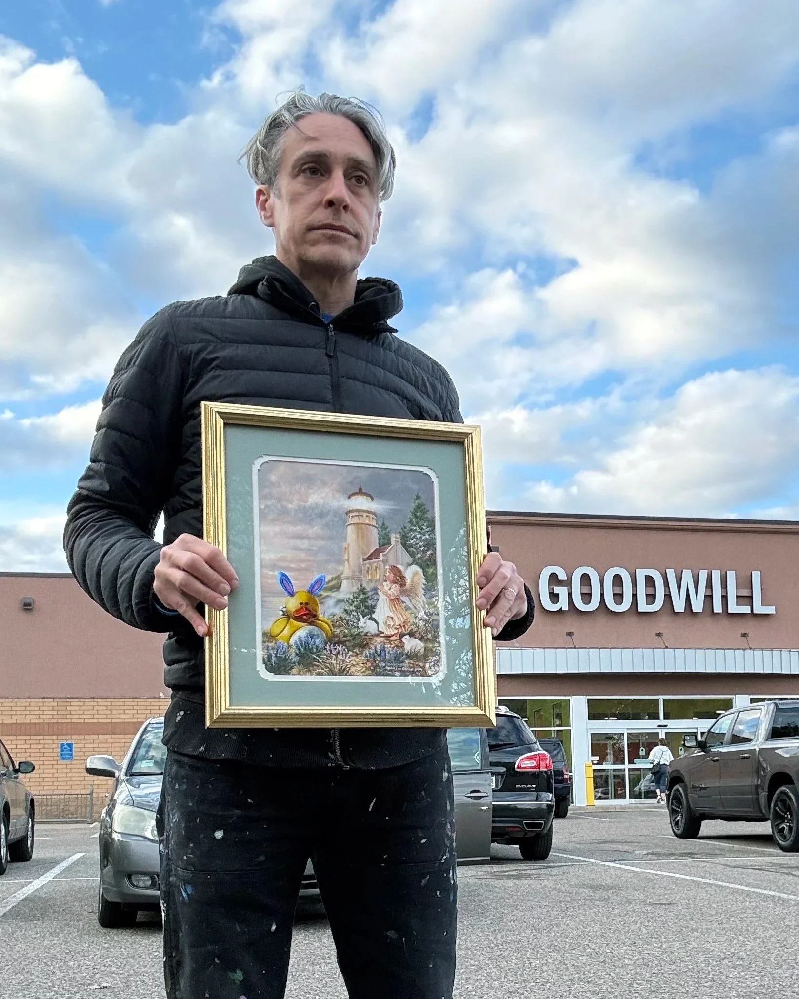 Man with a black coat holding a framed painting of an angel and a rubber duck in front of a Goodwill store.