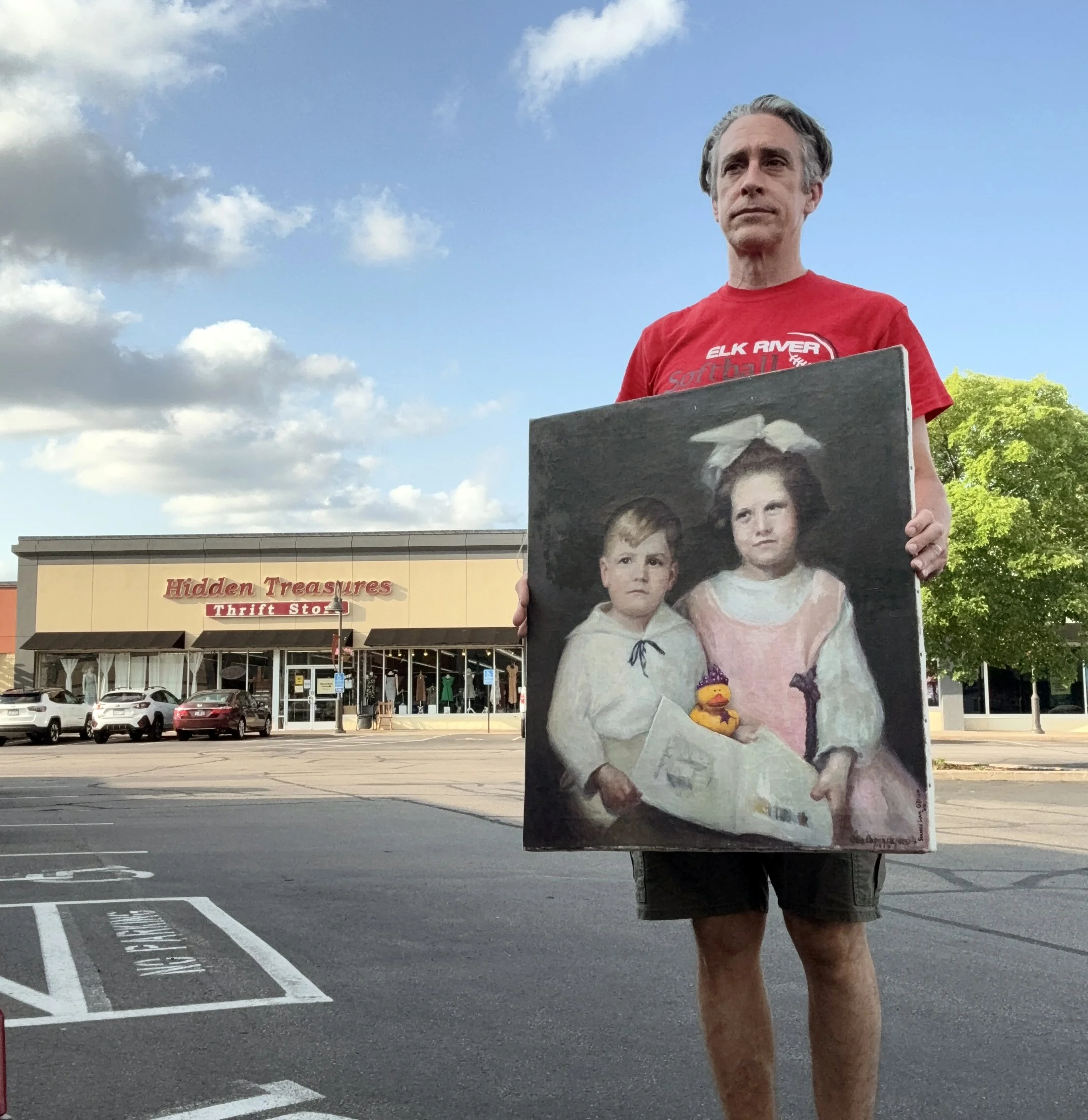 A man wearing a red shirt holding a portrait painting of two children and their yellow rubber duck in the parking lot of a thrift store.
