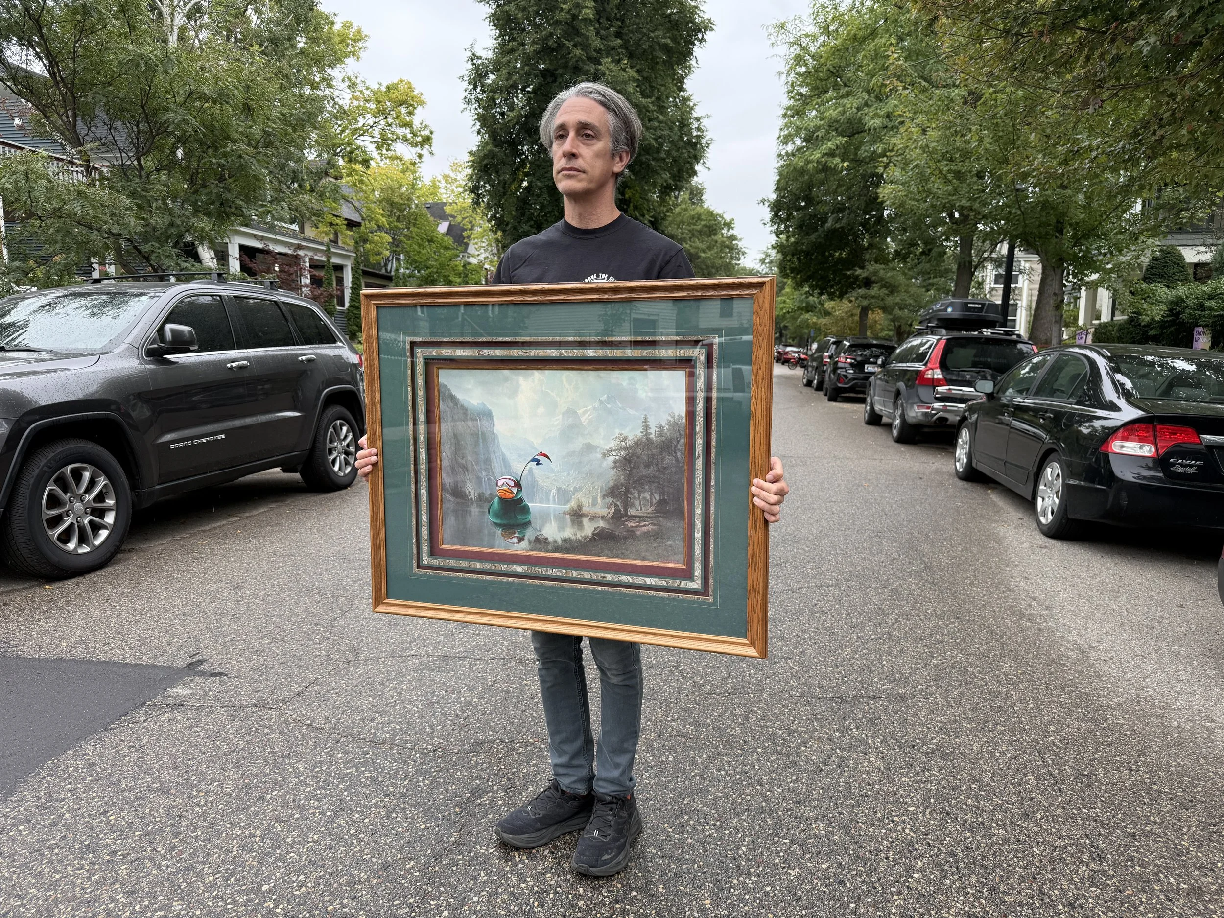 A man with big hair standing on the street holding a large framed landscape painting with a green rubber duck floating in a lake.