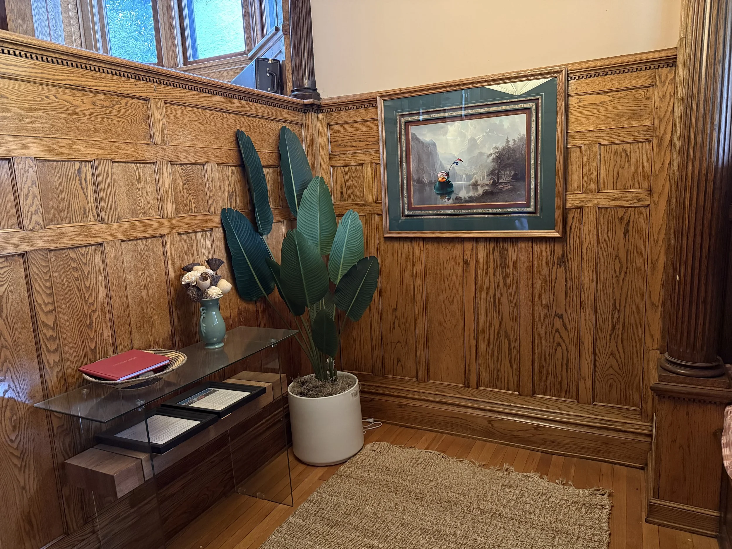 A green framed landscape painting hanging on a wooden wall next to a large green plant.