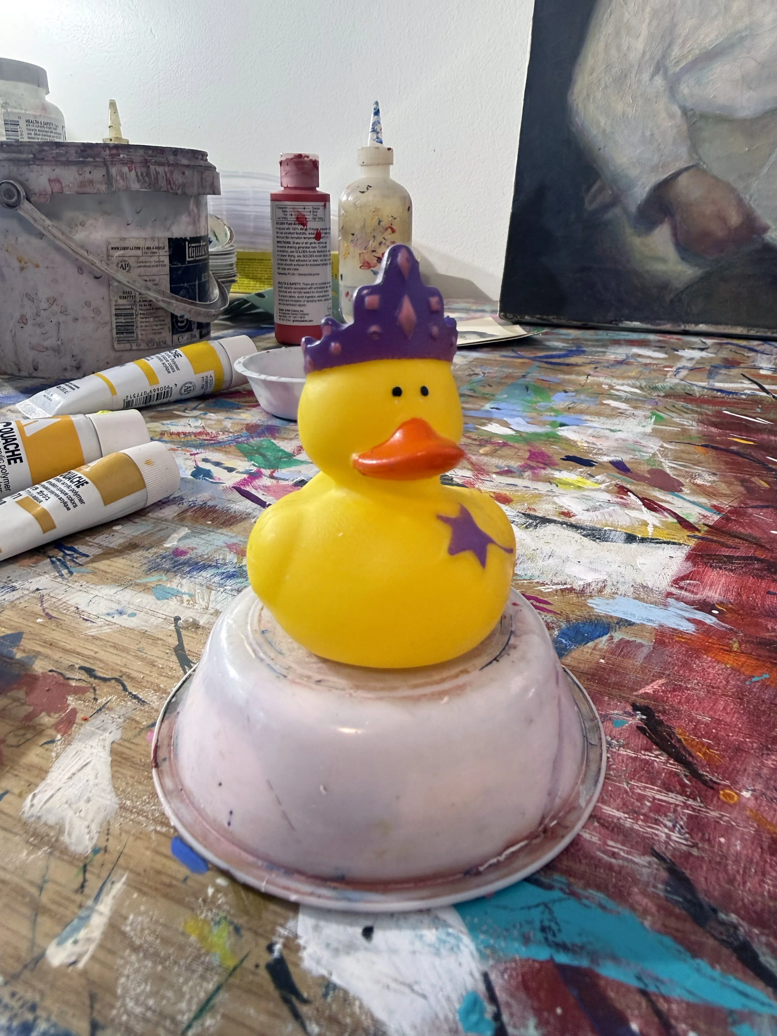 Yellow rubber duck toy wearing a purple crown sitting on a small cup on a painter's table.