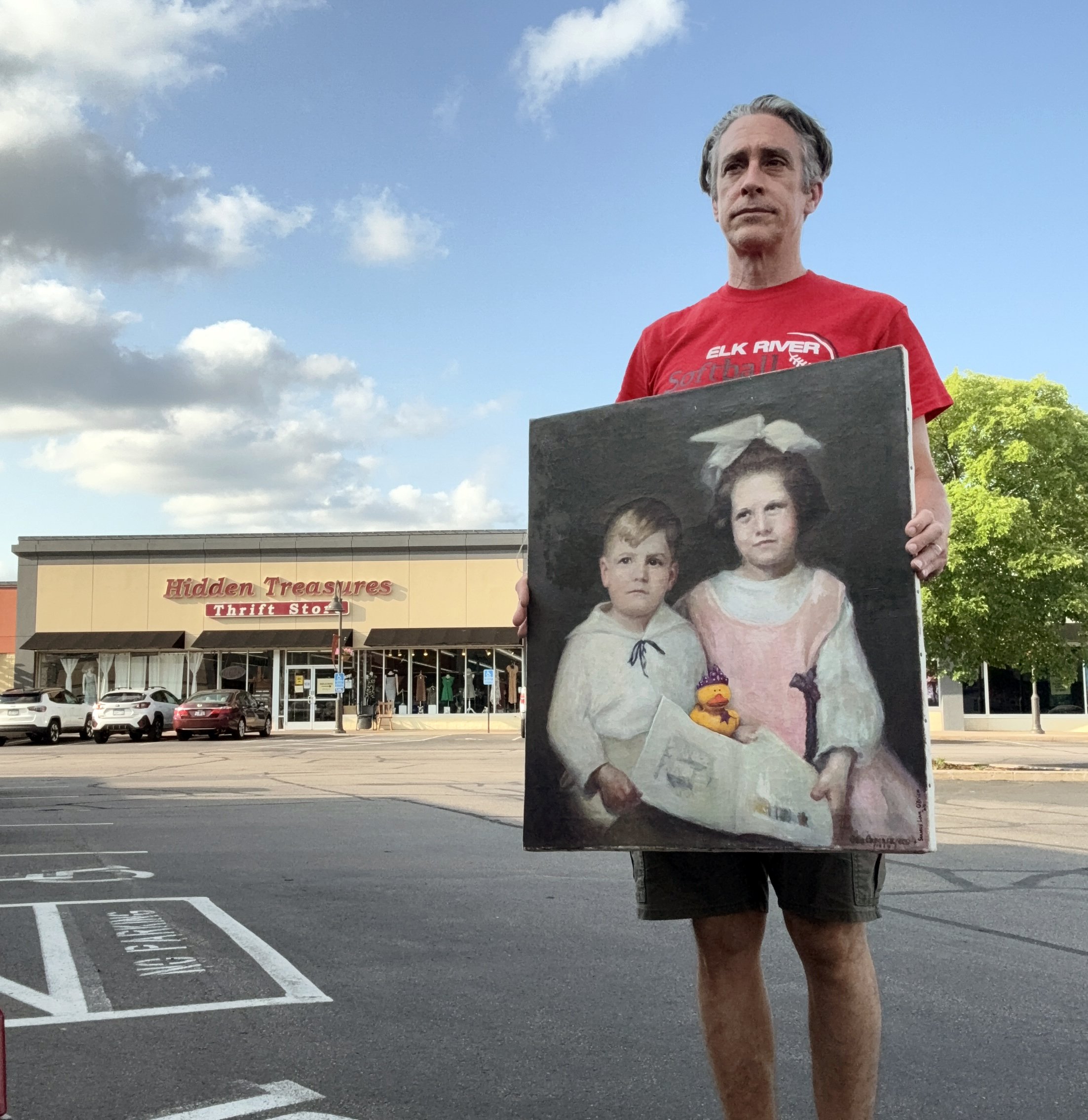 Man in a red shirt standing in a parking lot holding a portrait painting of two ghostly children.