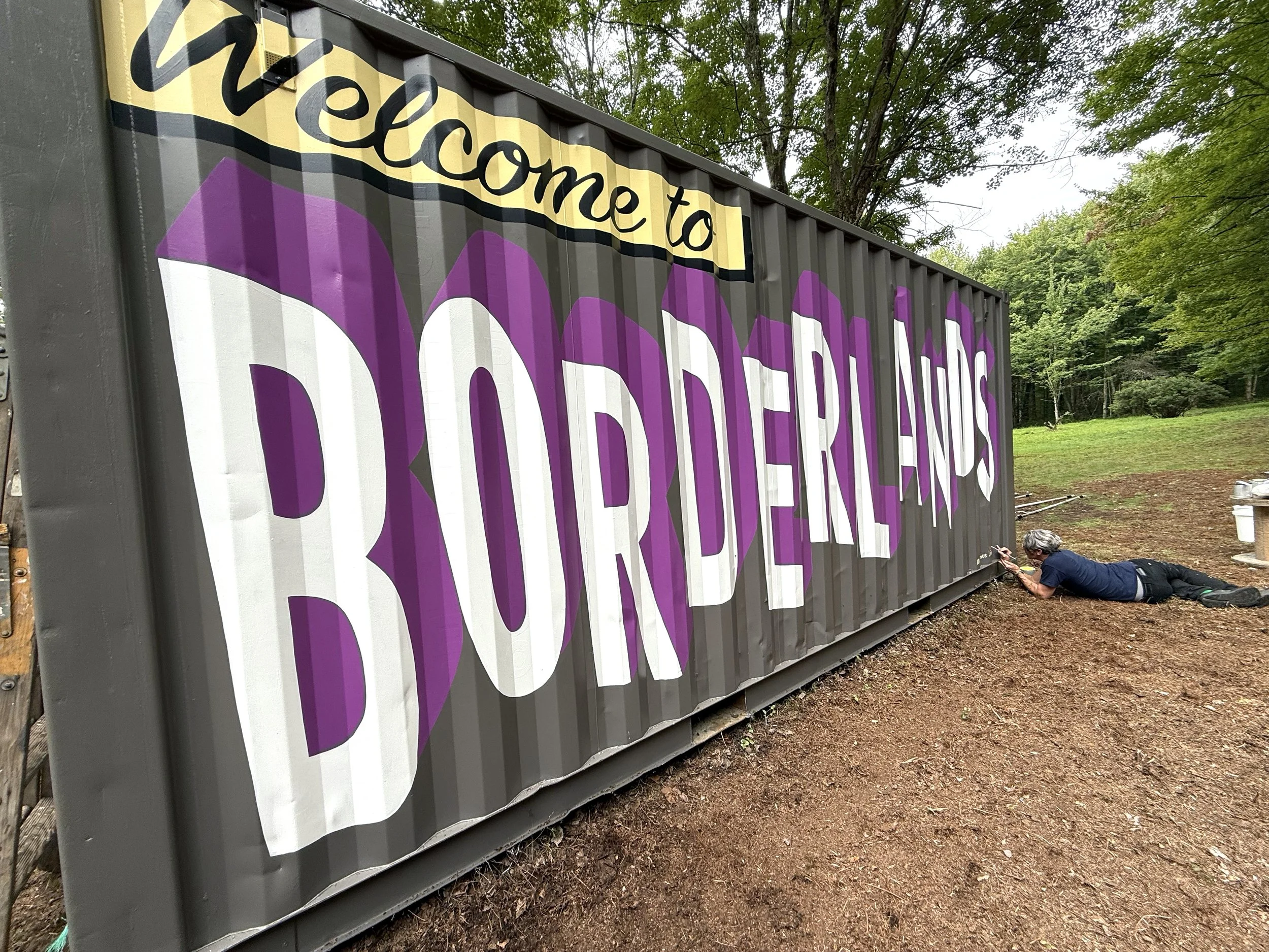 Man laying on the ground and painting some words on a grey shipping container