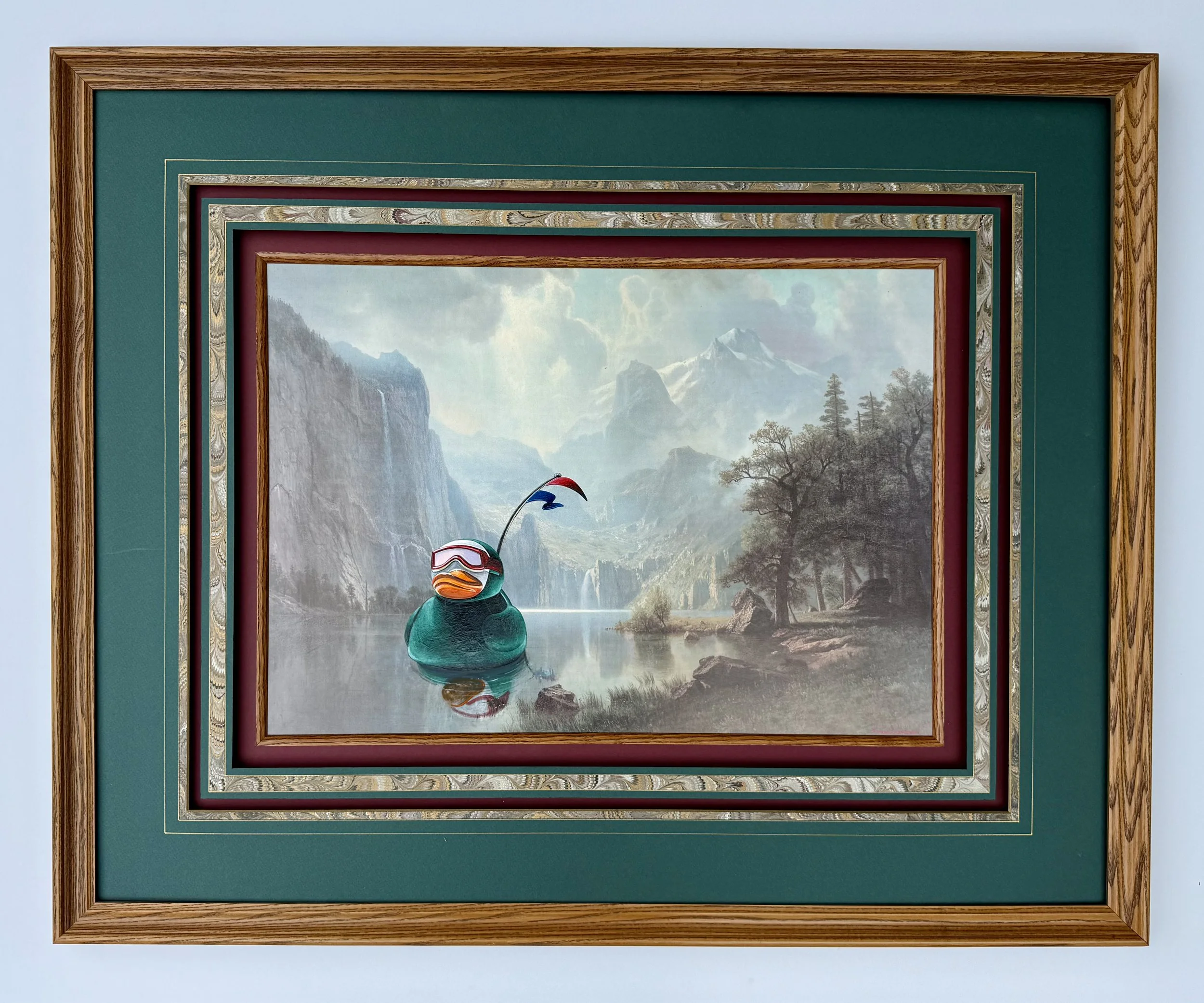 A framed painting of a large green rubber duck with goggles floating in a lake surrounded by mountains and wilderness.