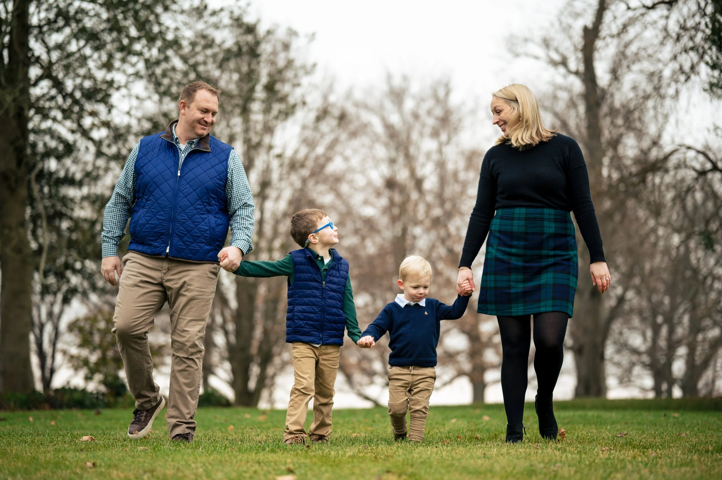 A family of four walking hand in hand through a park on a cloudy day, with trees in the background.