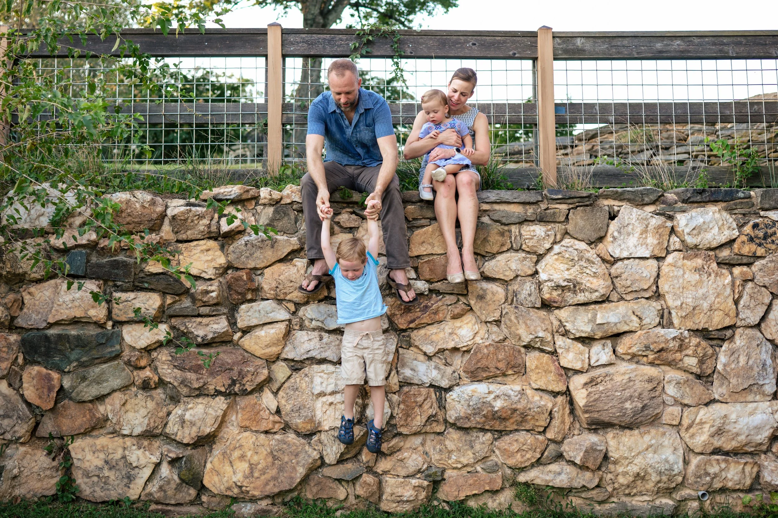 A family outdoors with a rocky wall; a woman holds a young girl, a man is helping a young boy climb the wall, who is holding the man's hands.