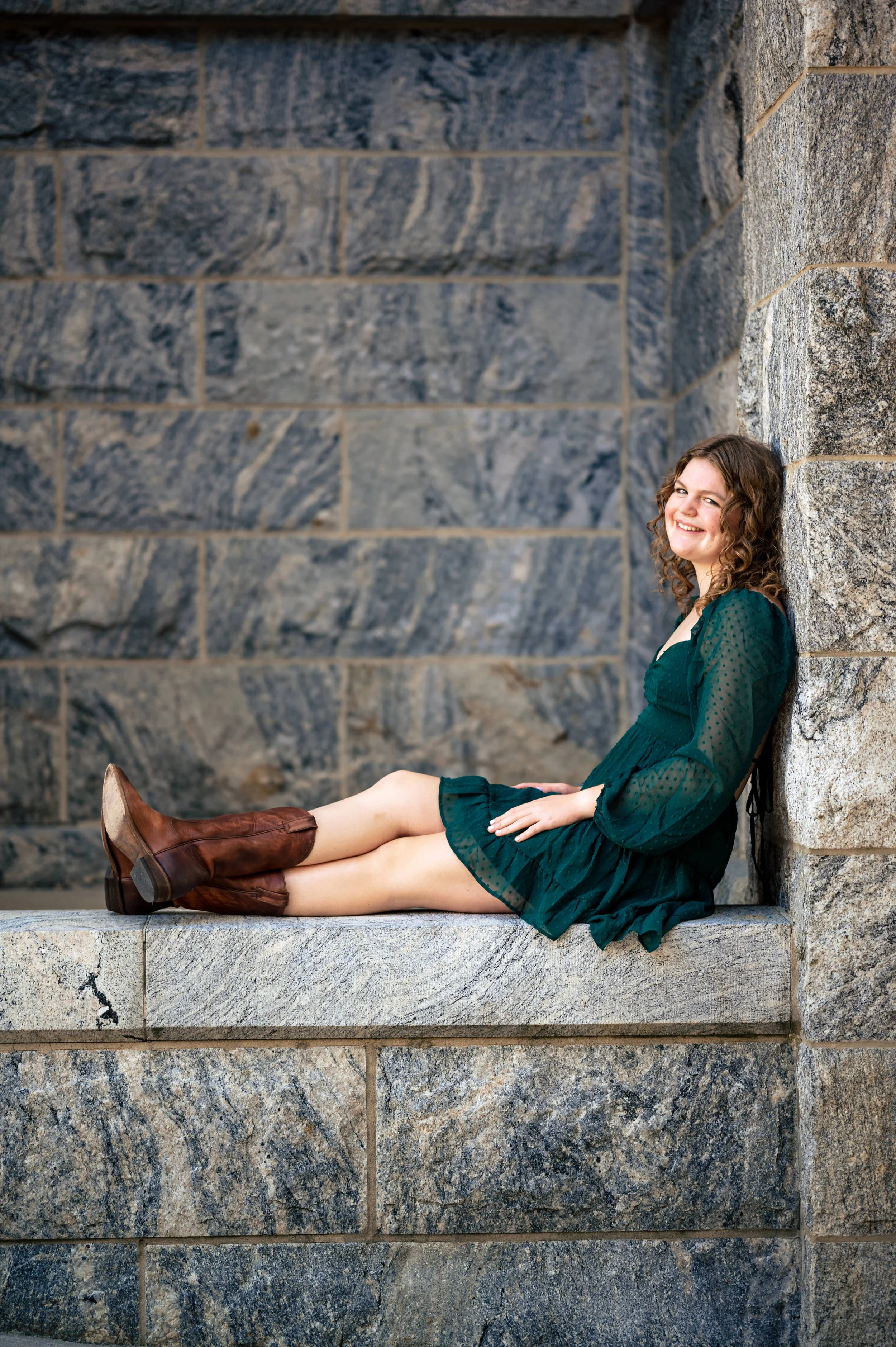 A smiling young woman with curly hair, wearing a green dress and brown boots, sitting on a stone ledge against a stone wall.