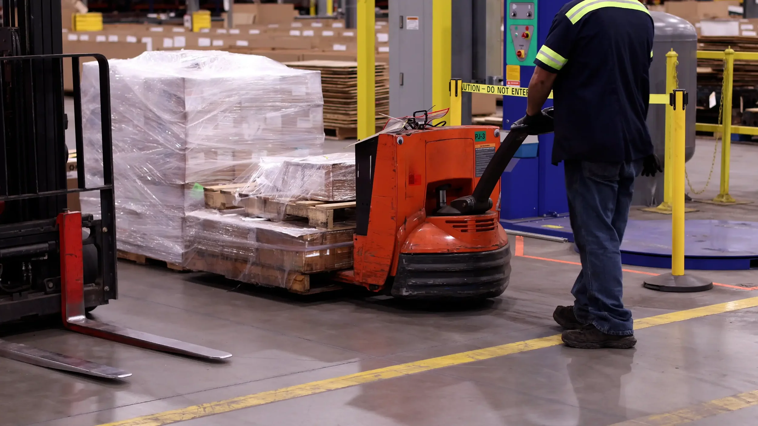 Warehouse worker operating pallet jack to move a pallet wrapped in plastic containing boxes, with shelving and safety barriers in the background.